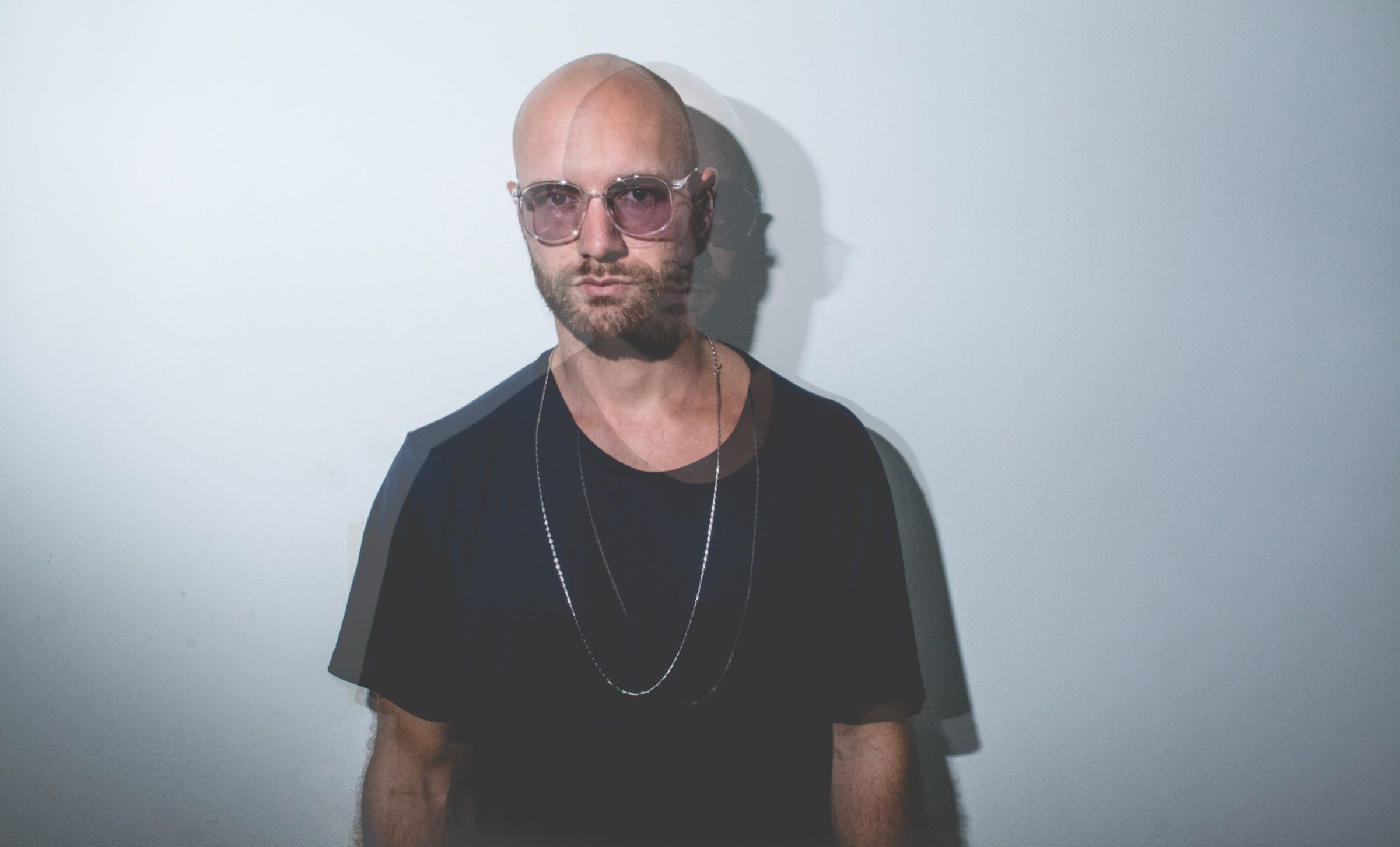 Double-exposure portrait of a bald man with a beard, tinted glasses and a chain necklace staring into the camera against a pale wall
