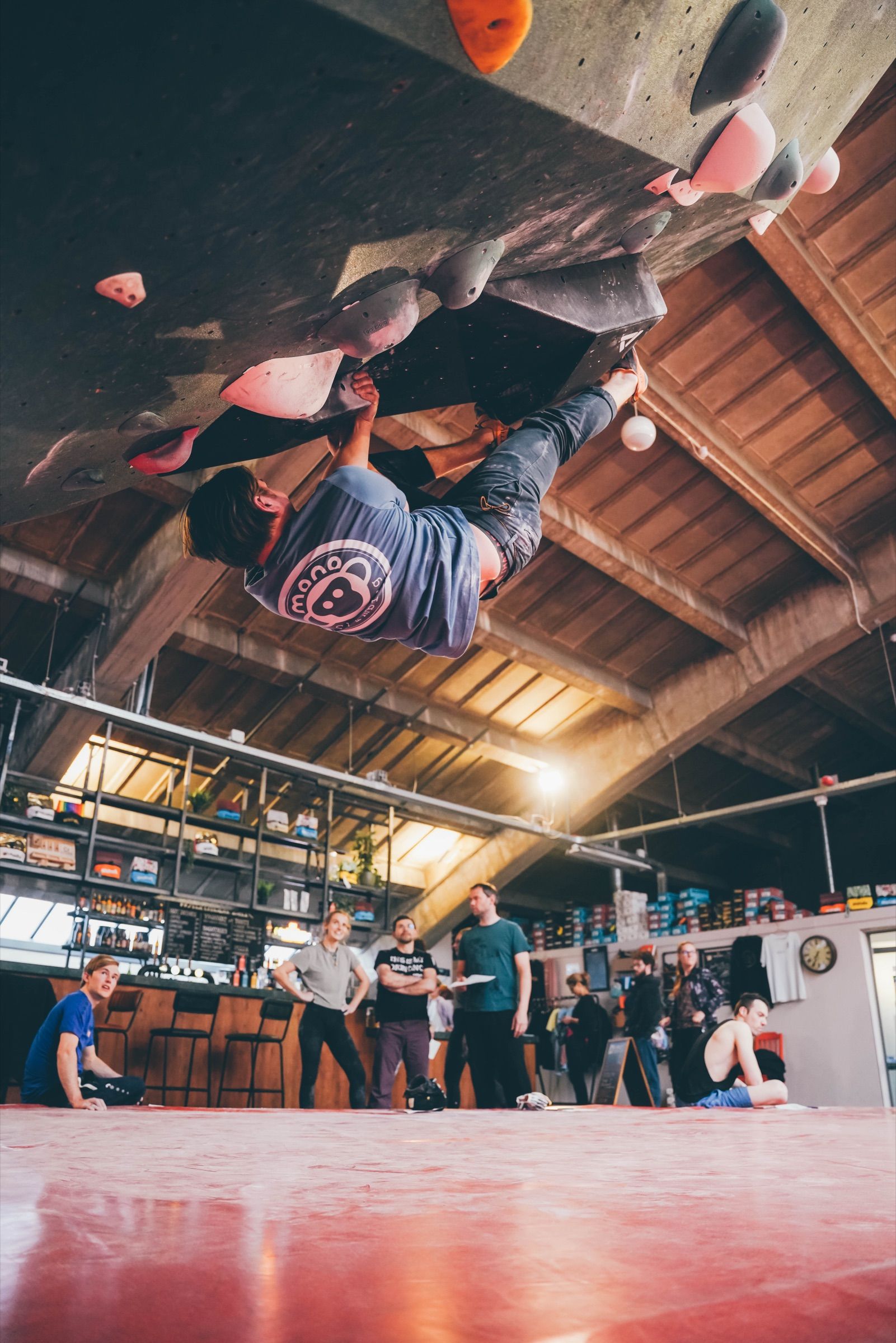 Climber hanging upside down beneath the overhanging roof of an indoor bouldering wall while onlookers watch from the gym bar below