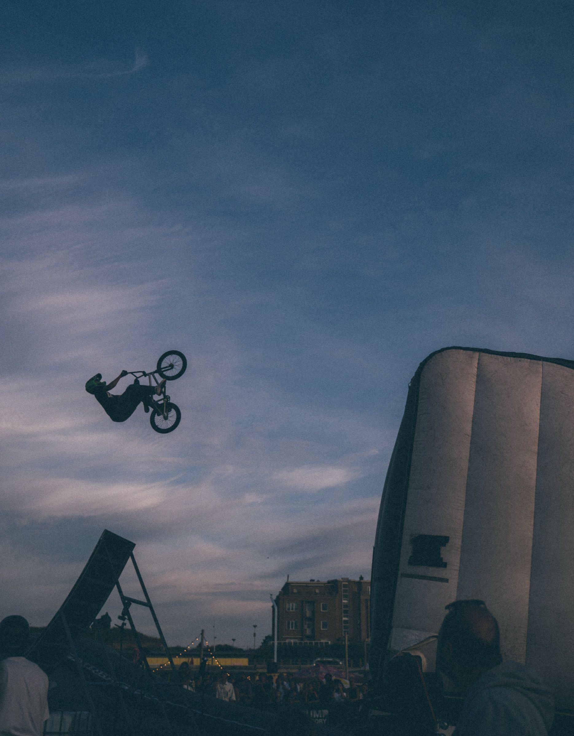 BMX rider silhouetted upside down against a dusky evening sky between a launch ramp and an inflatable landing airbag