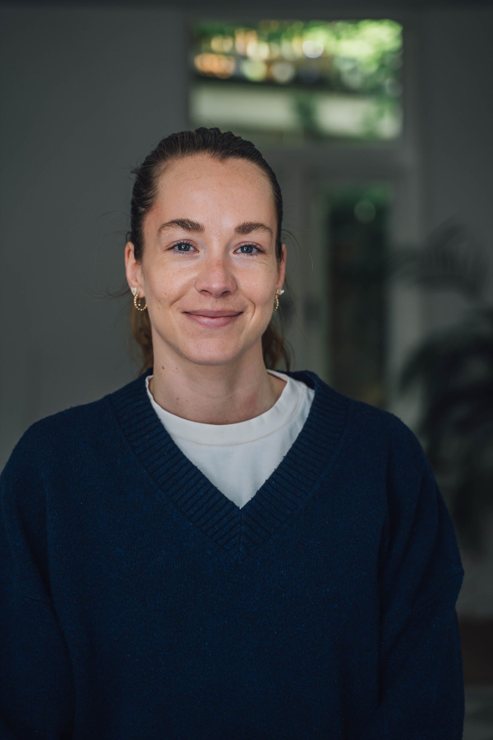 Portrait of a woman with her hair tied back and gold hoop earrings, in a navy V-neck jumper over a white top, smiling softly by a bright window