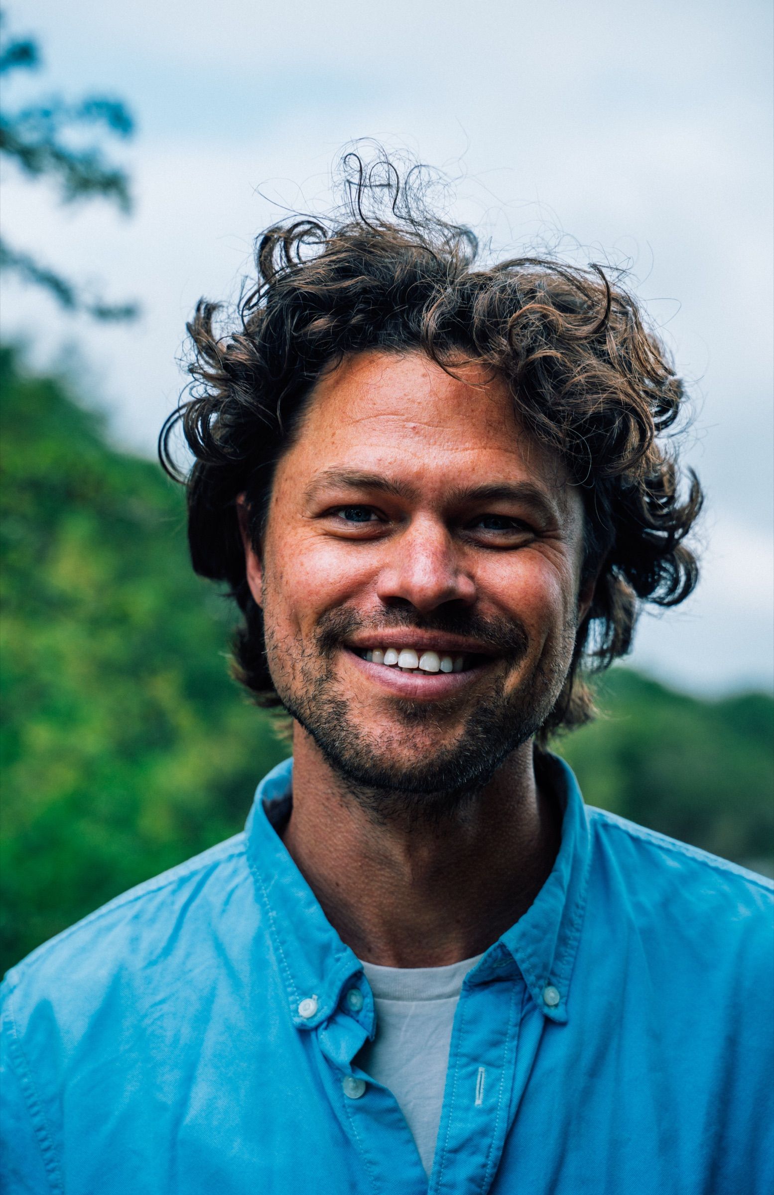 Outdoor portrait of a curly-haired man with a broad, genuine smile in a blue button-up shirt, green foliage and soft sky behind him