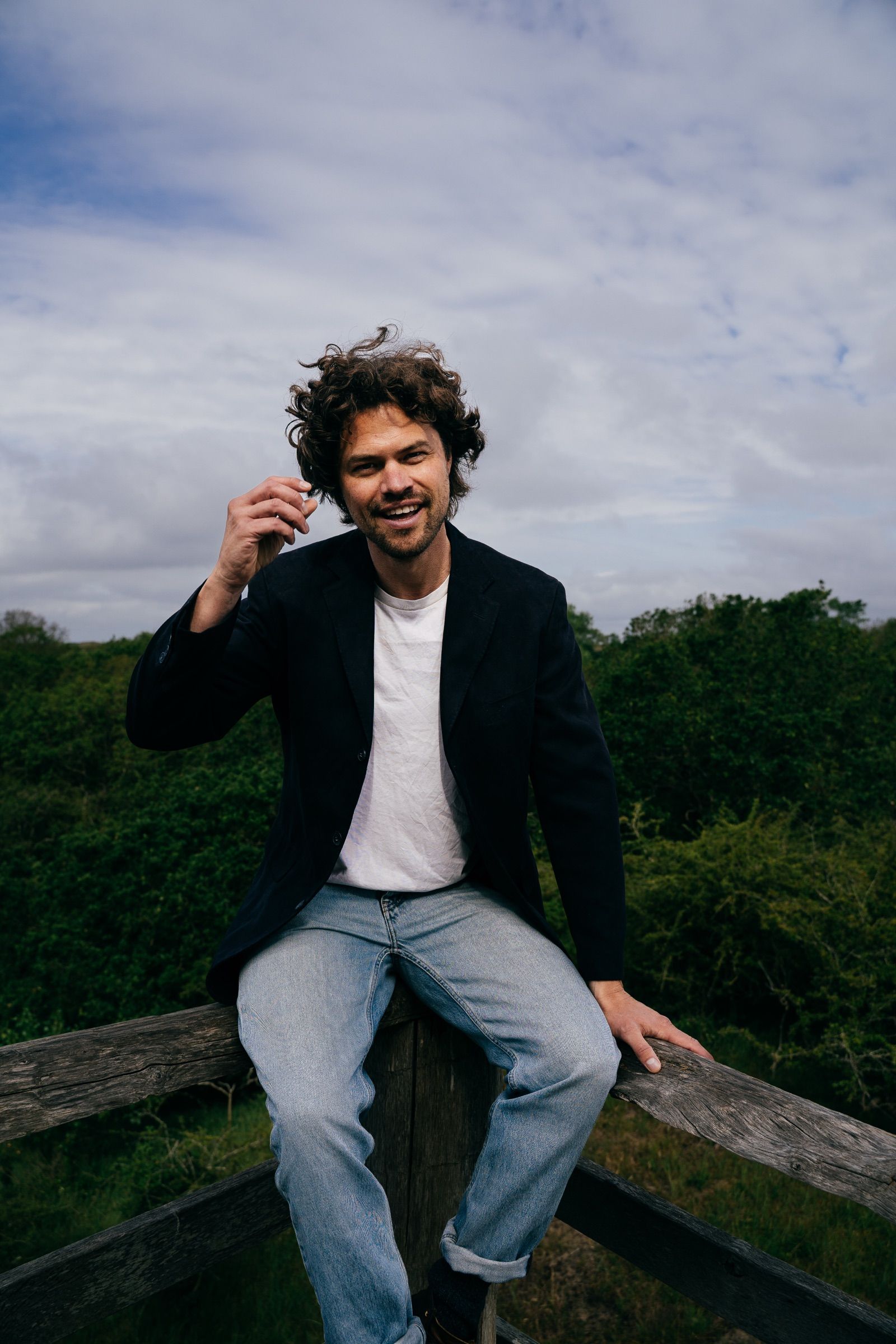 Man in a navy blazer, white tee and jeans laughing while perched on a weathered wooden fence, a cloudy sky over green dunes behind him