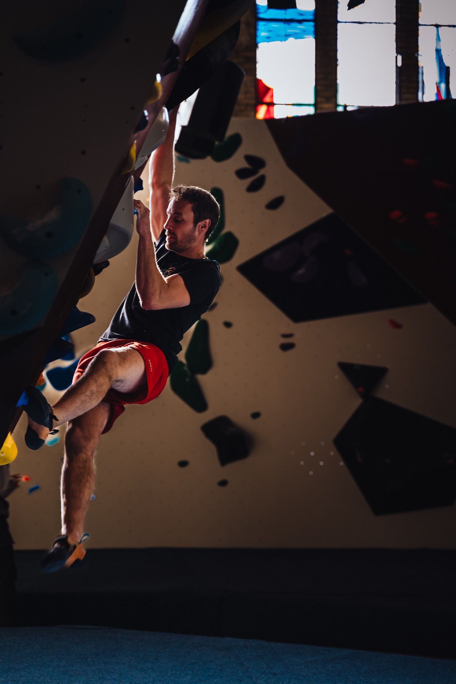 Climber in red shorts hanging one-armed from an overhanging bouldering wall, backlit by a bright gym window