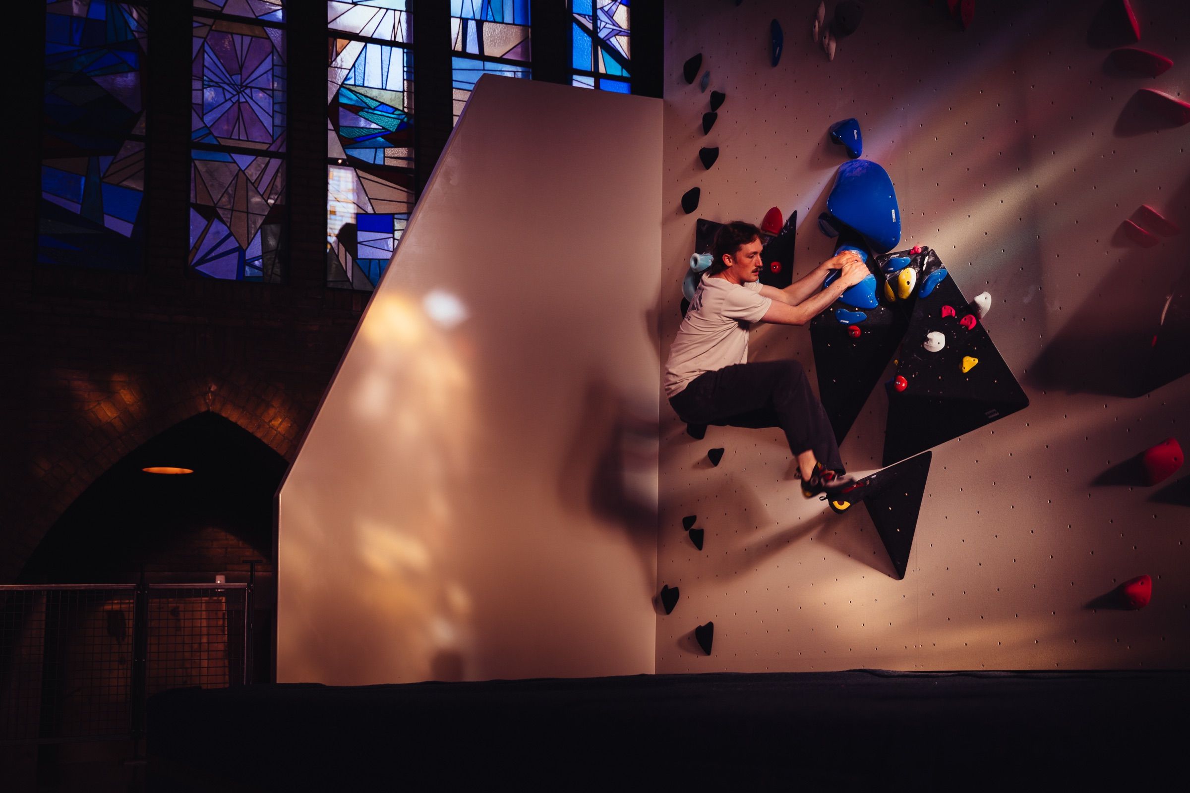 Climber in a white t-shirt working up black angular volumes on a bouldering wall inside a converted church with stained-glass windows