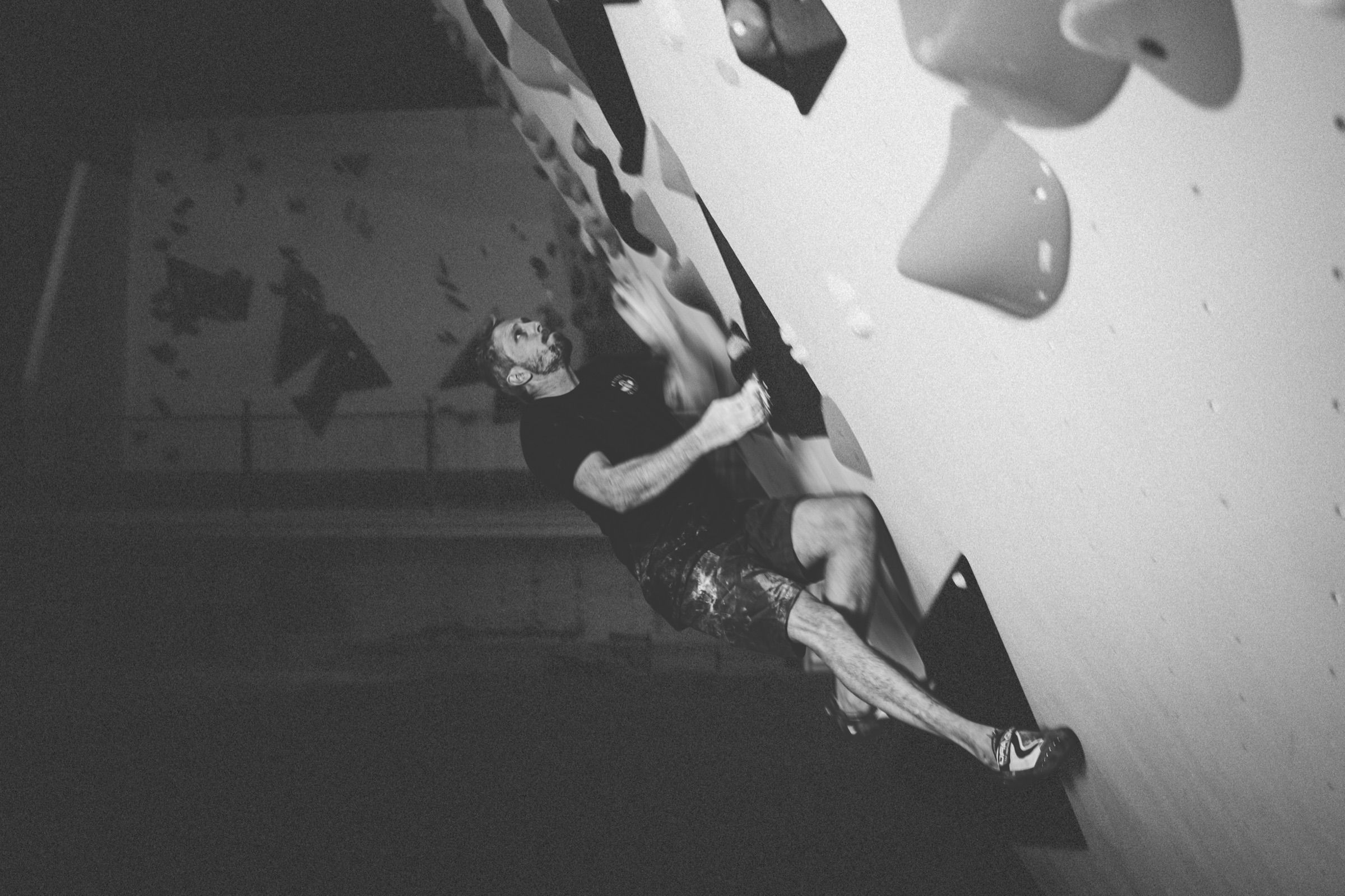 Tilted black-and-white view of a boulderer in a tank top reaching between large holds on a pale overhanging gym wall