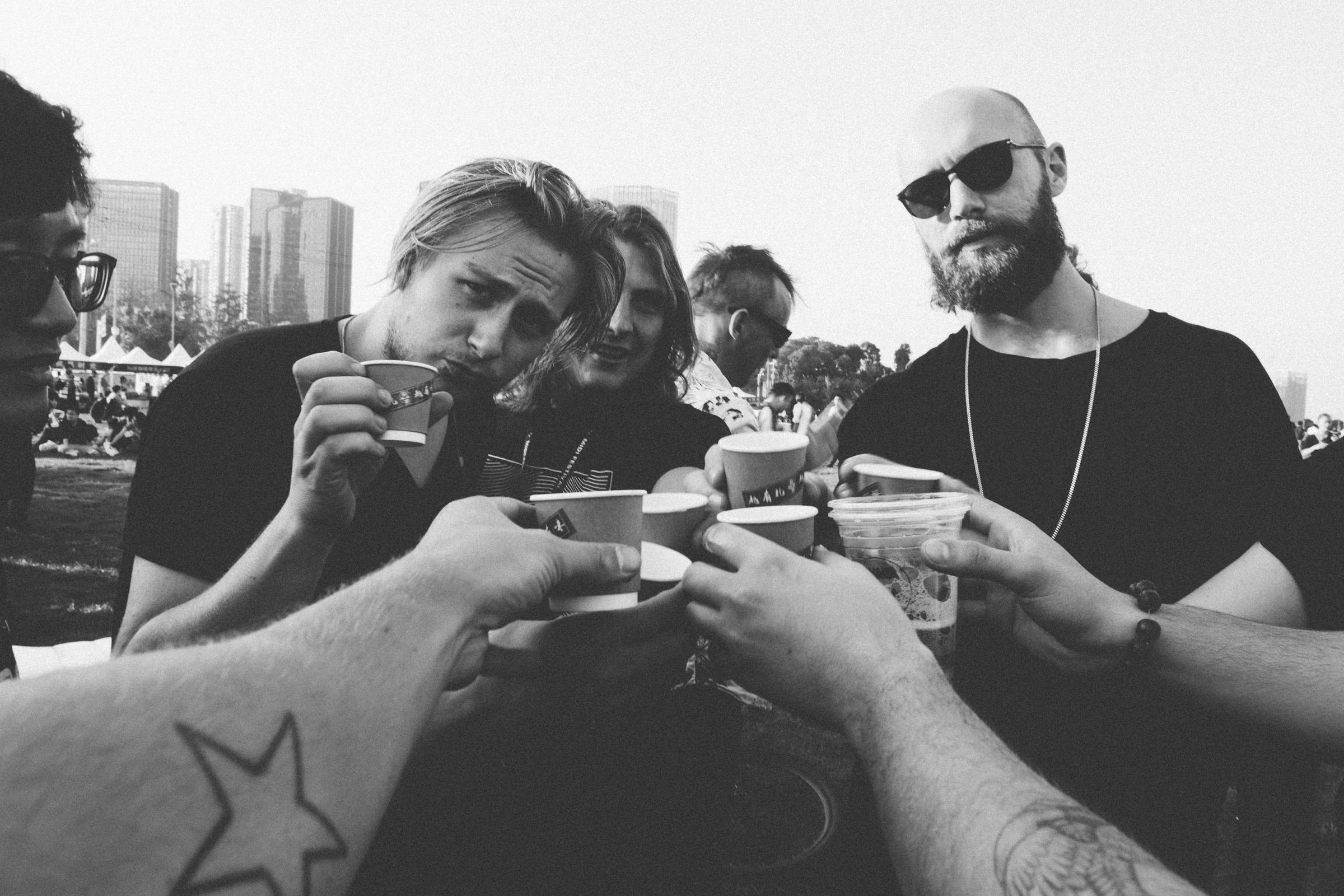 Black-and-white moment of friends leaning in to toast with paper cups and a beer at an outdoor festival, tattooed forearms in the foreground