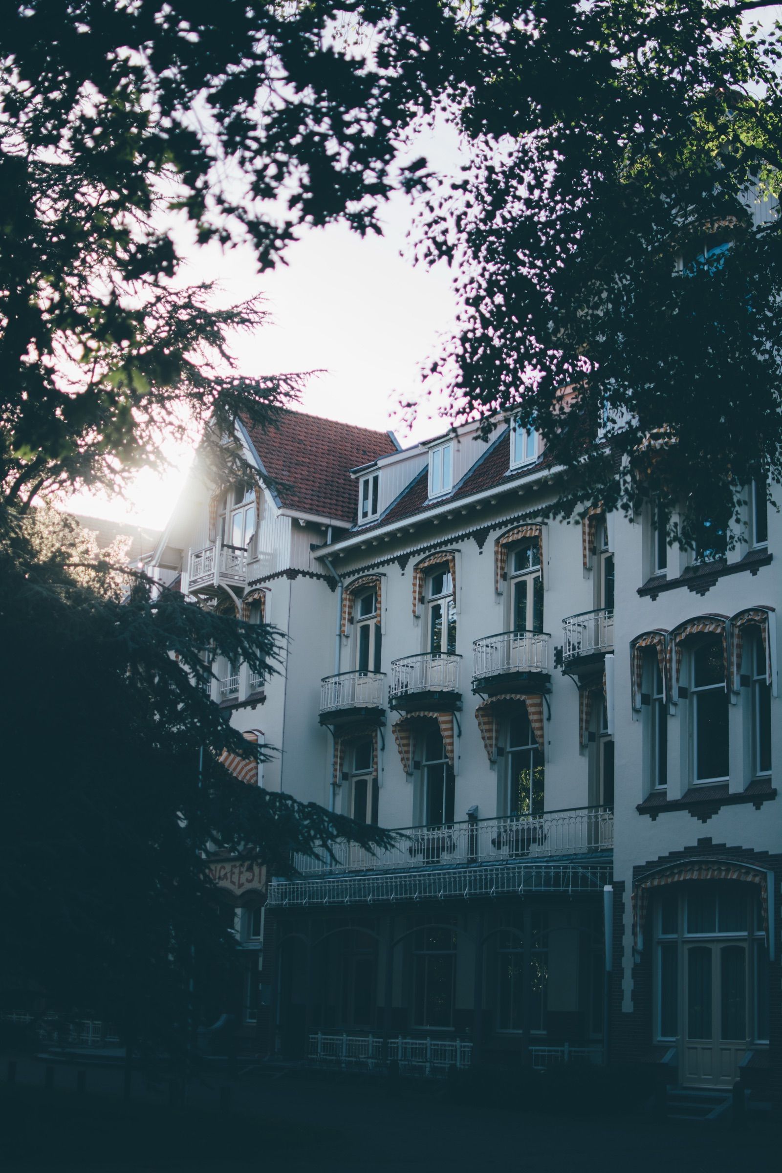 Stately white residence with balconies, striped awnings and a red tiled roof glimpsed through dark tree branches in low evening sun