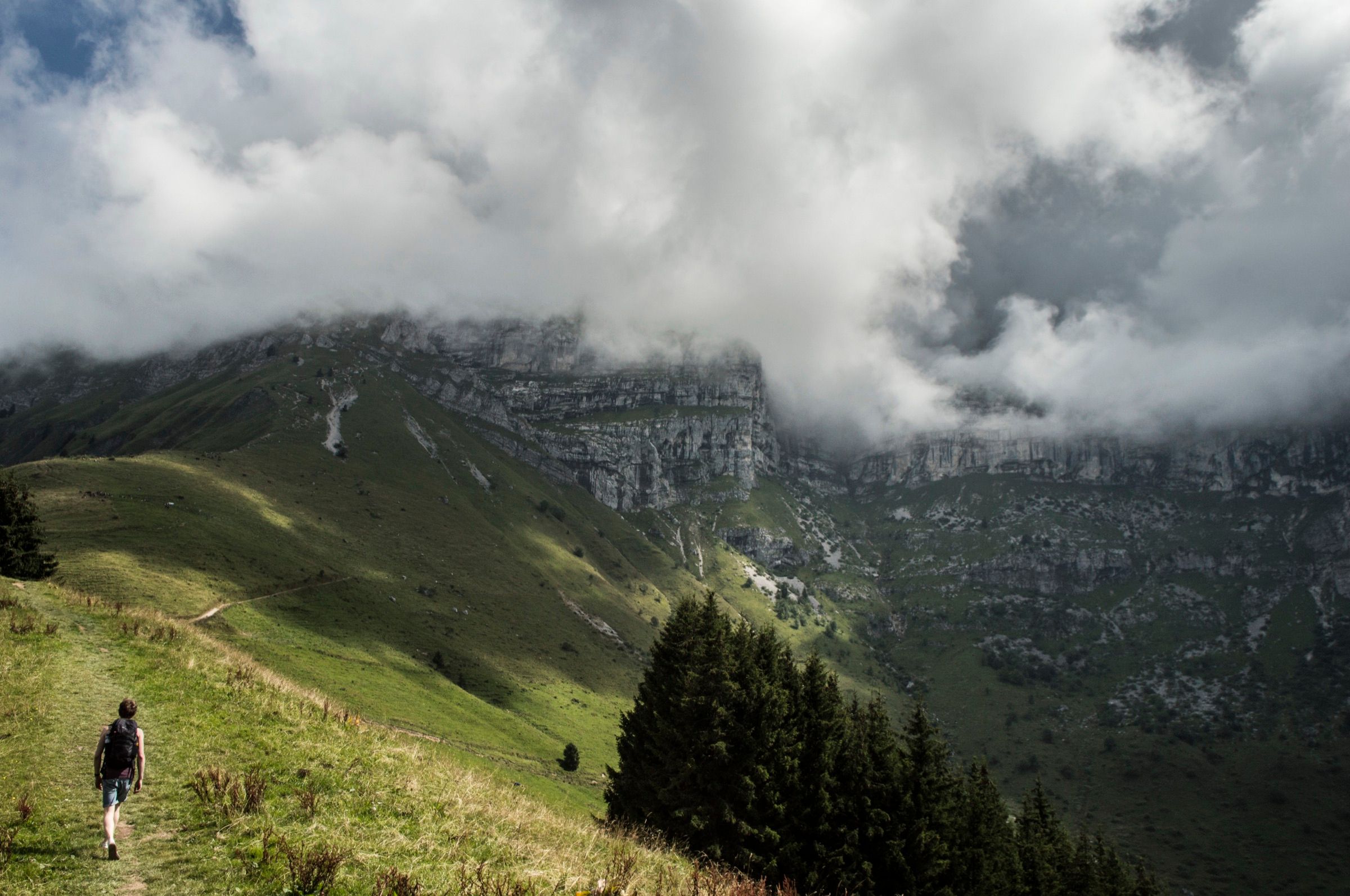 Hiker with a backpack following a grassy ridge path toward limestone cliffs wrapped in rolling clouds