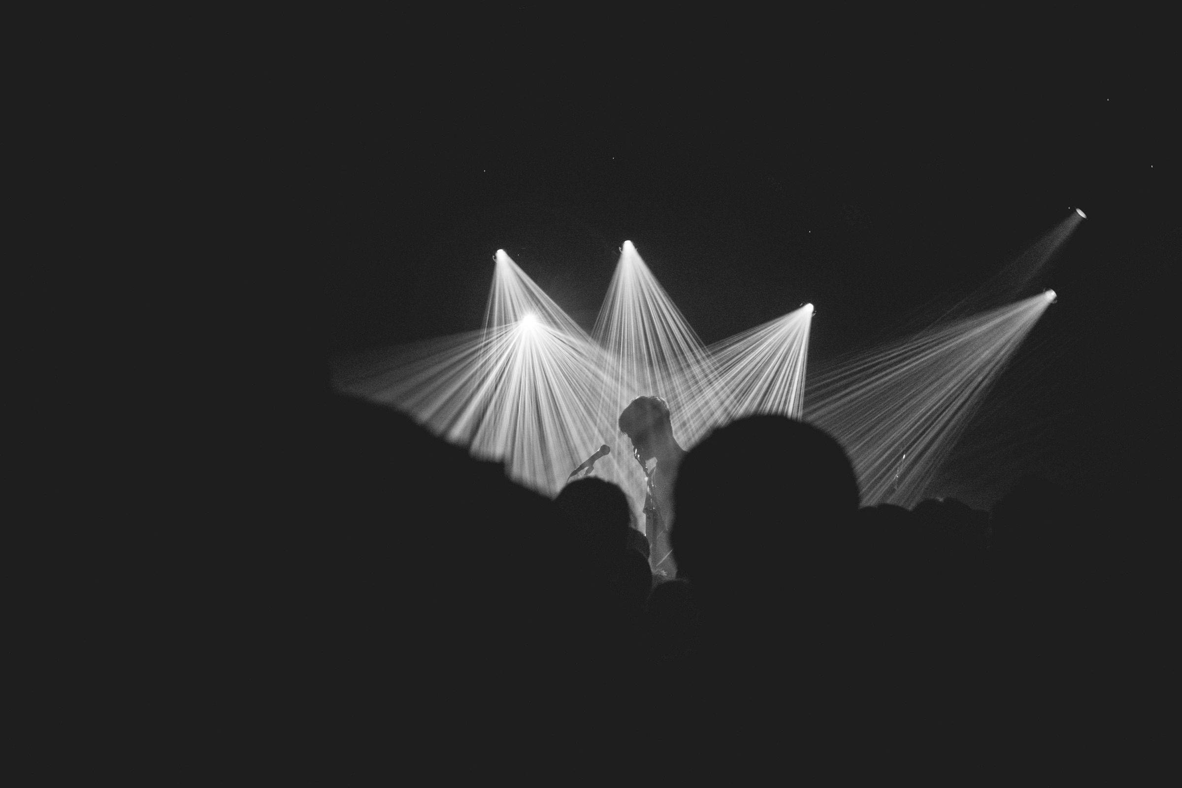 Black-and-white silhouette of a singer at a microphone beneath a fan of white spotlight beams above a dark audience