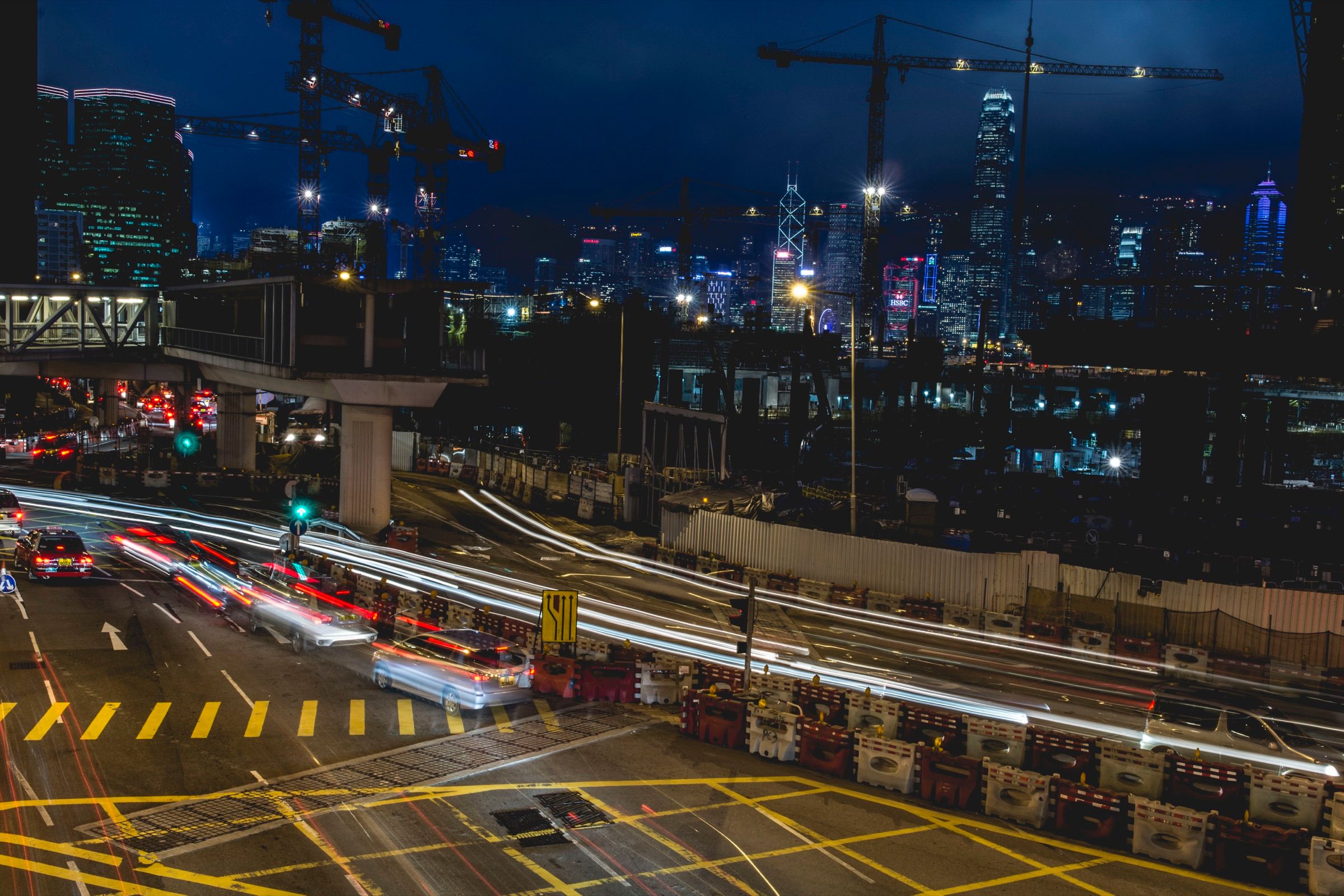 Long-exposure night view of traffic light trails sweeping through a construction-lined intersection beneath a glowing high-rise skyline