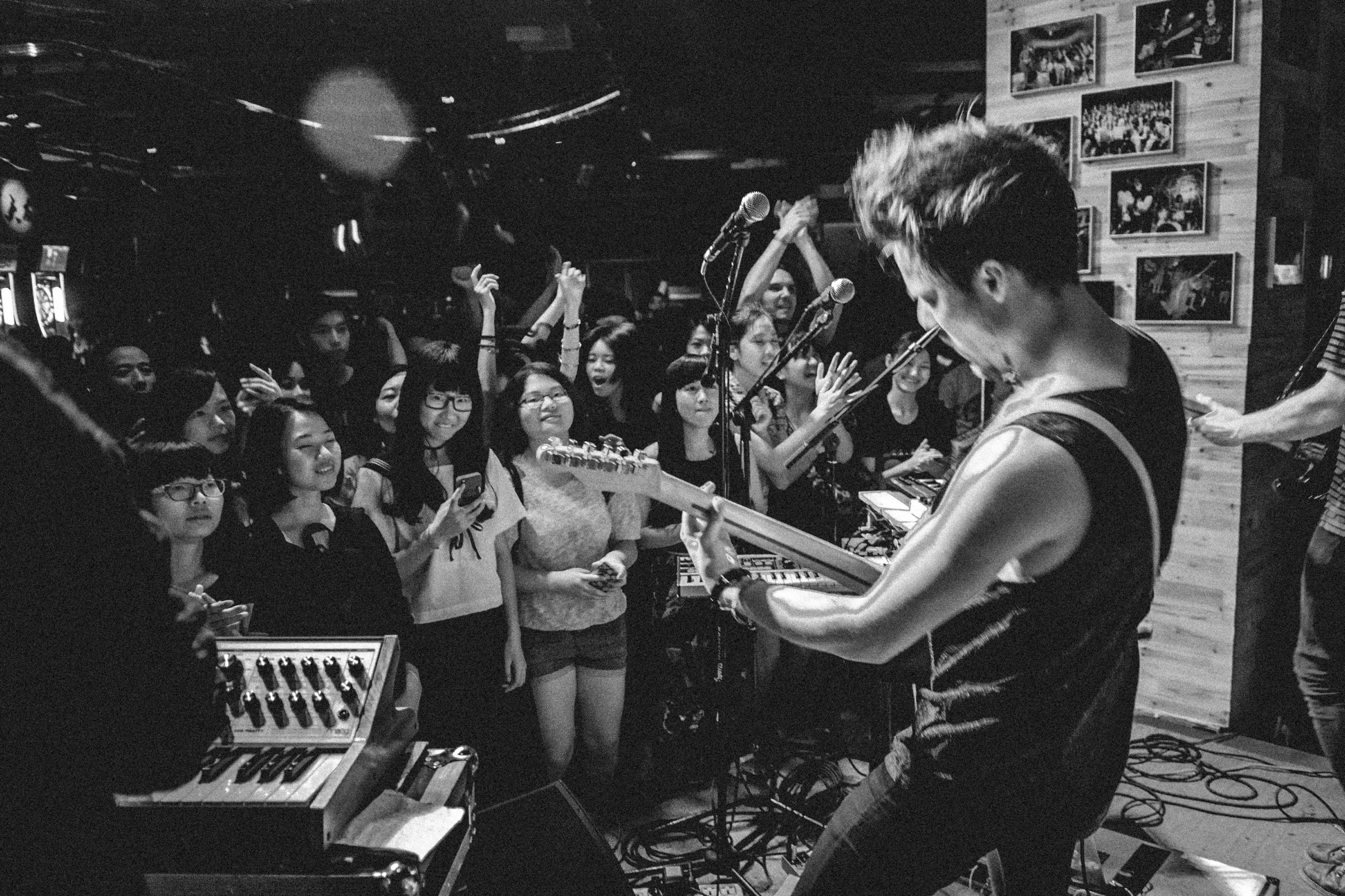 Black-and-white view over a guitarist’s shoulder of a packed club crowd smiling and clapping close to the stage