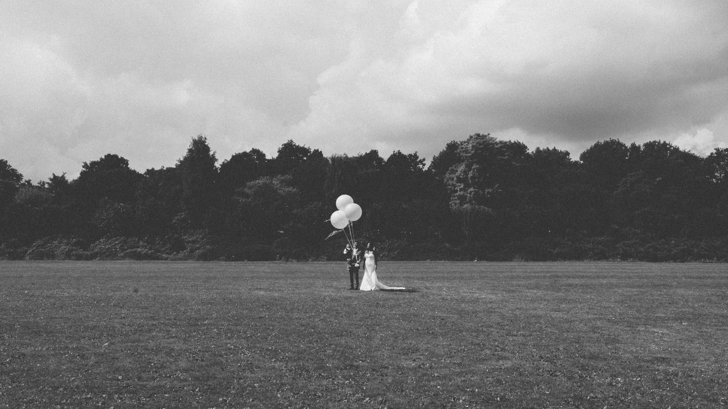 Black-and-white field scene with a bride and groom standing small in the distance holding a cluster of giant white balloons
