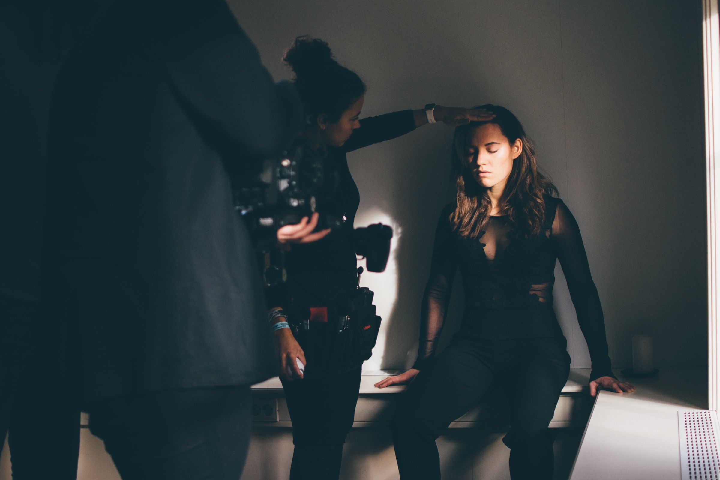 Stylist adjusting a seated model’s hair in a dark room where a shaft of window light falls across the model’s face