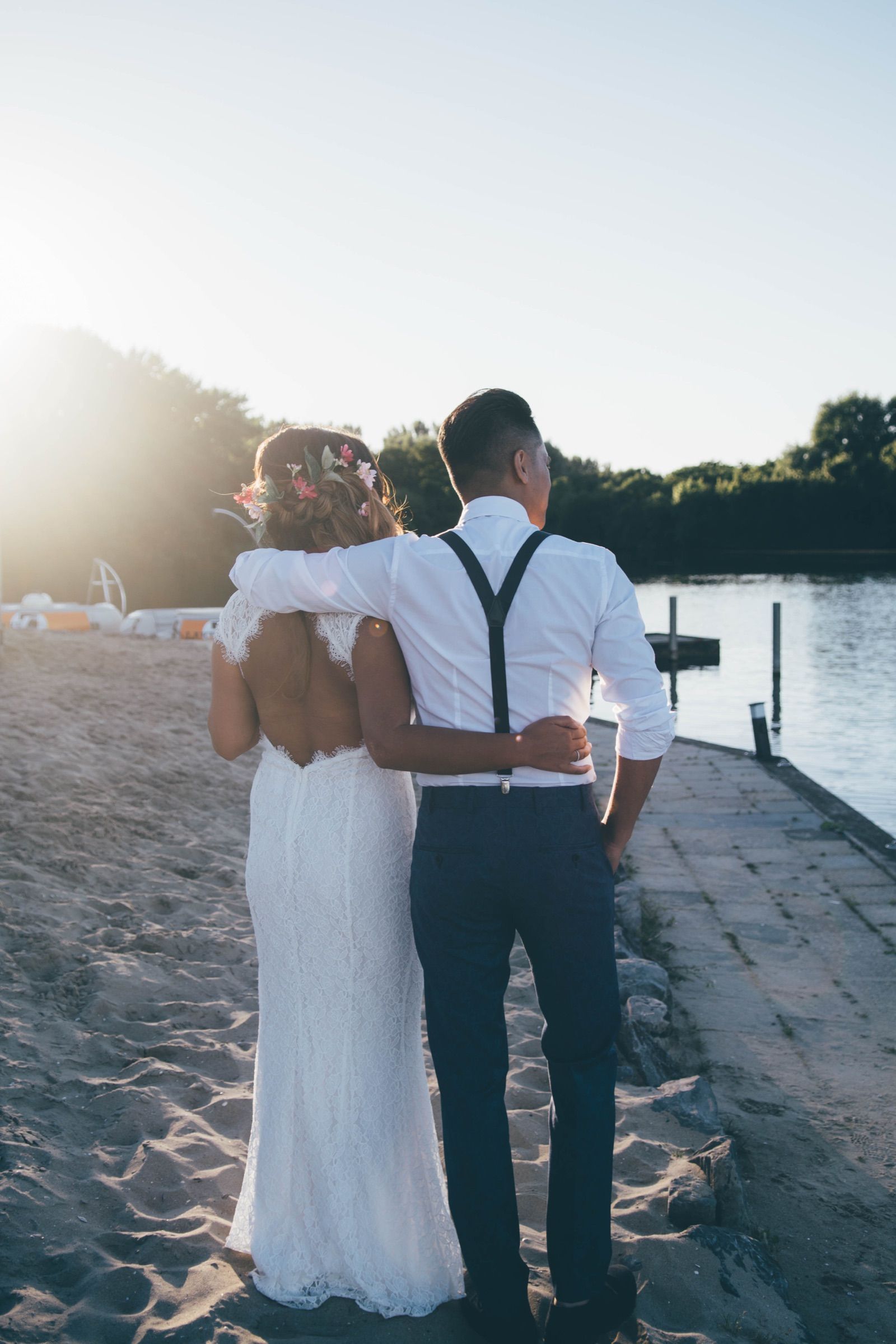 Bride with flowers in her braided hair and groom in suspenders standing arm in arm on lakeside sand in low golden light
