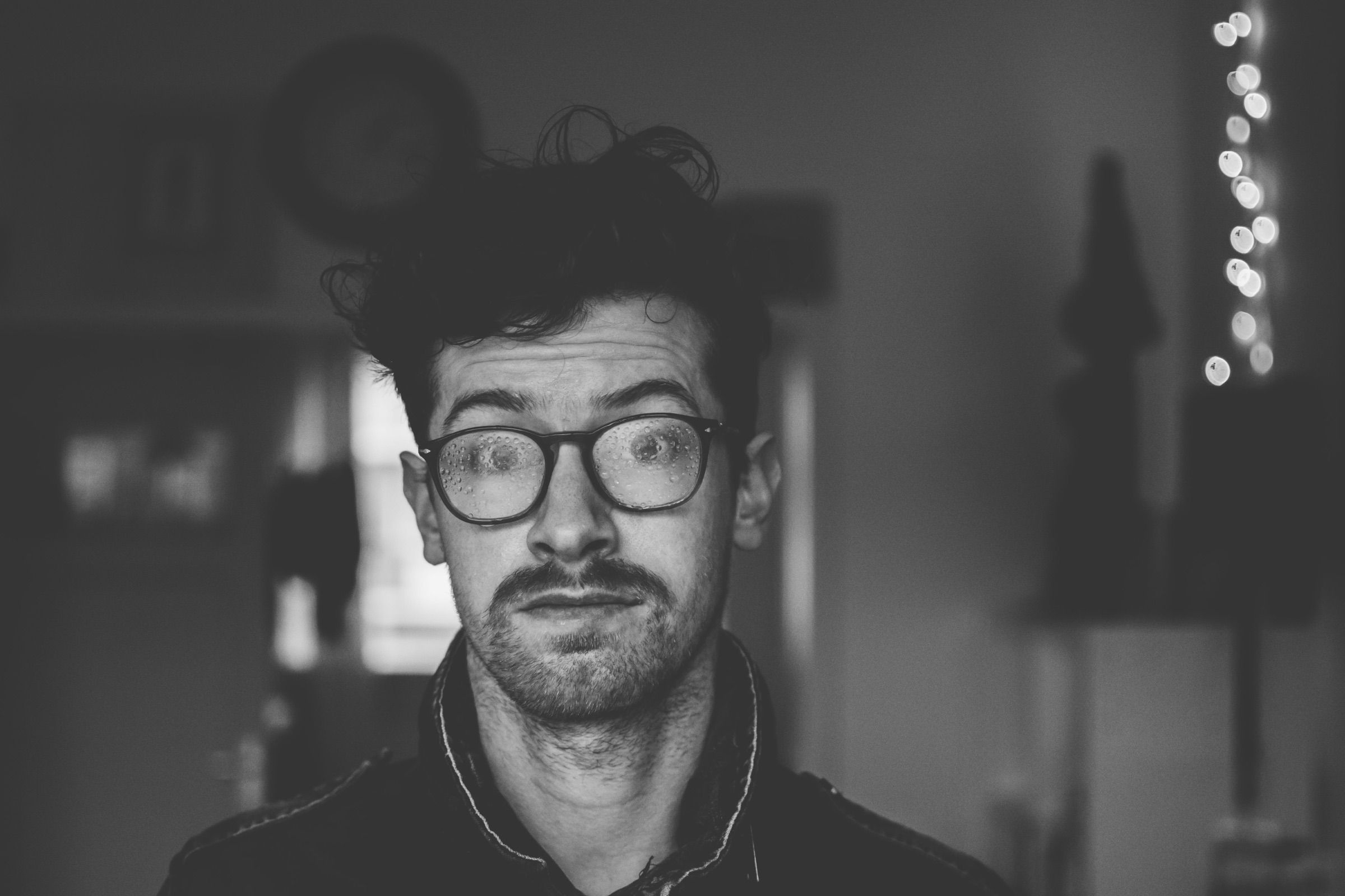 Black-and-white portrait of a man with tousled hair raising an eyebrow behind rain-fogged round glasses, string lights glowing behind