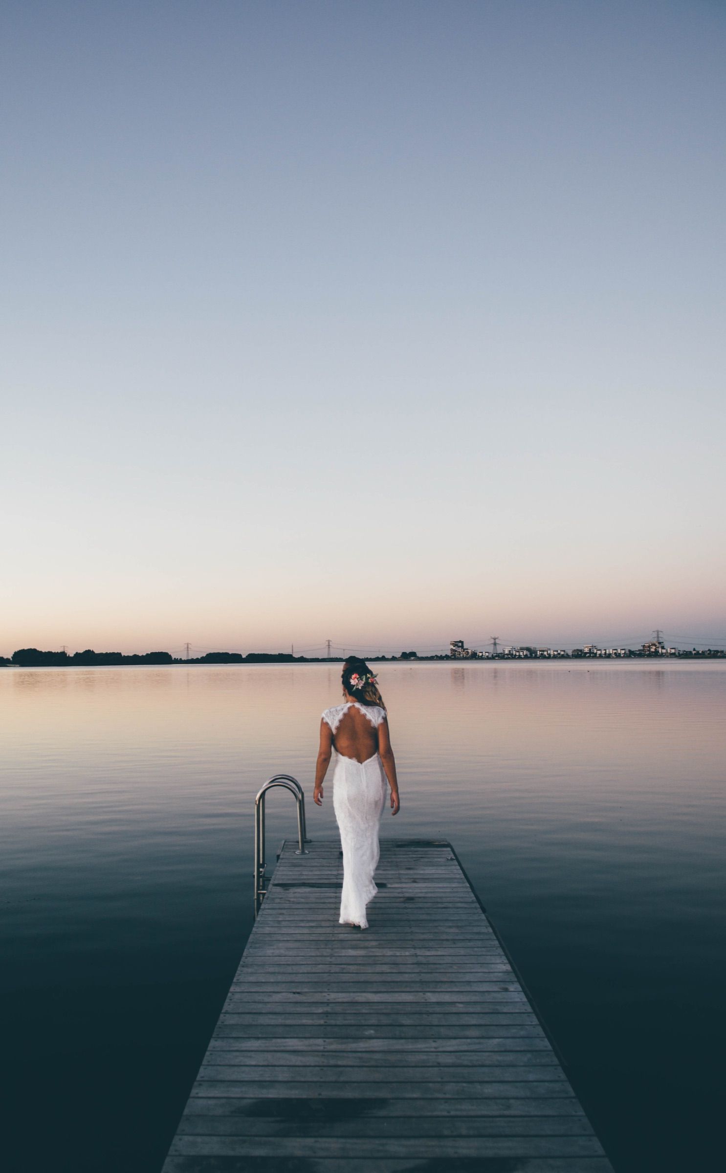Bride in a backless lace gown walking away down a wooden swimming jetty over a glass-calm lake at dusk