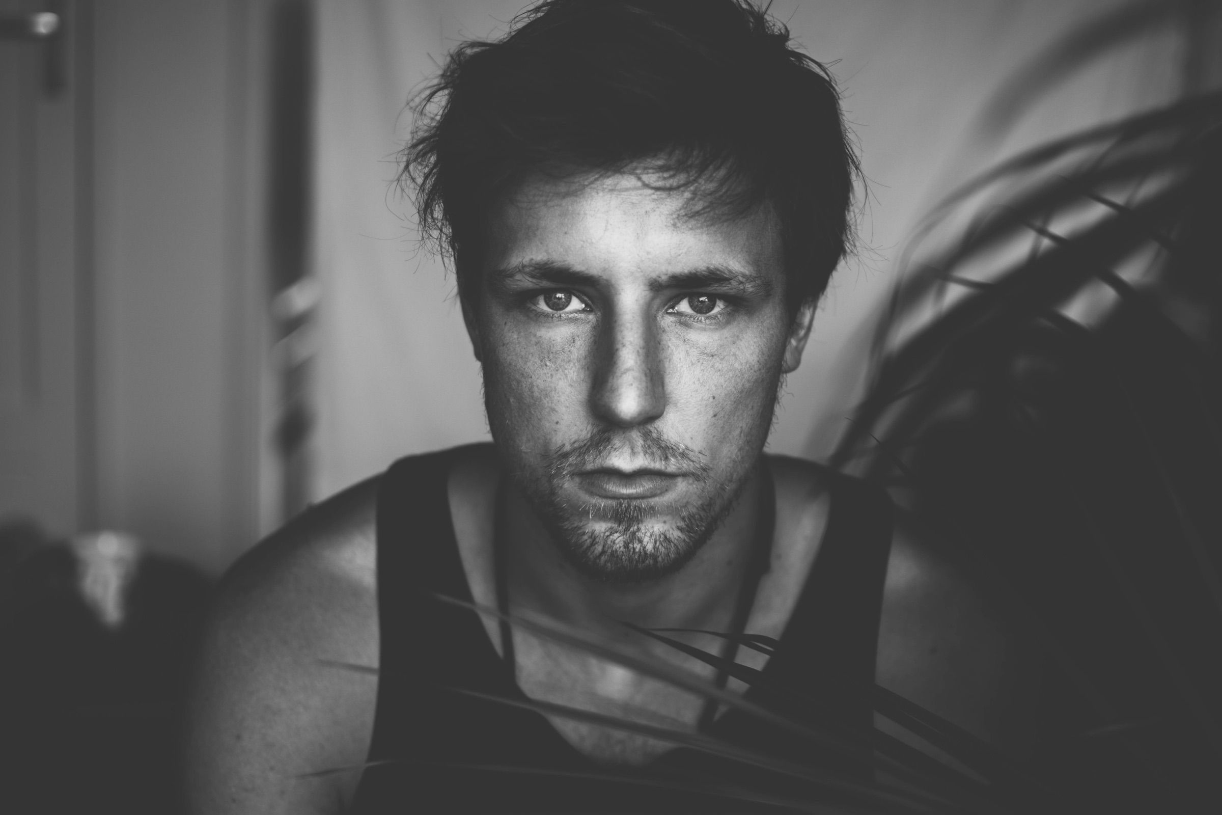 Black-and-white close portrait of a man in a tank top gazing steadily at the camera through drifting palm fronds