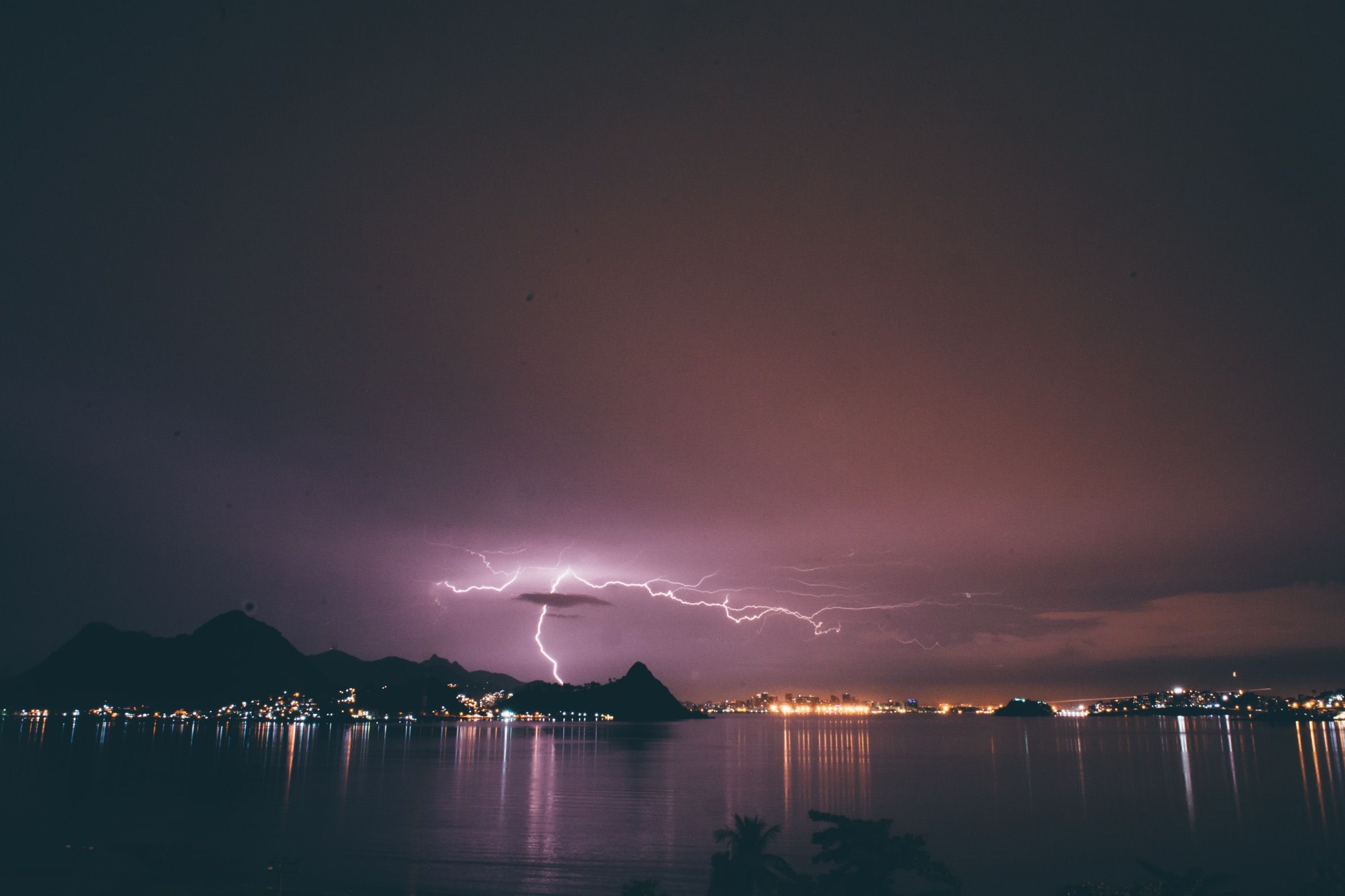 Forked lightning striking from a violet night sky over a bay lined with distant city lights