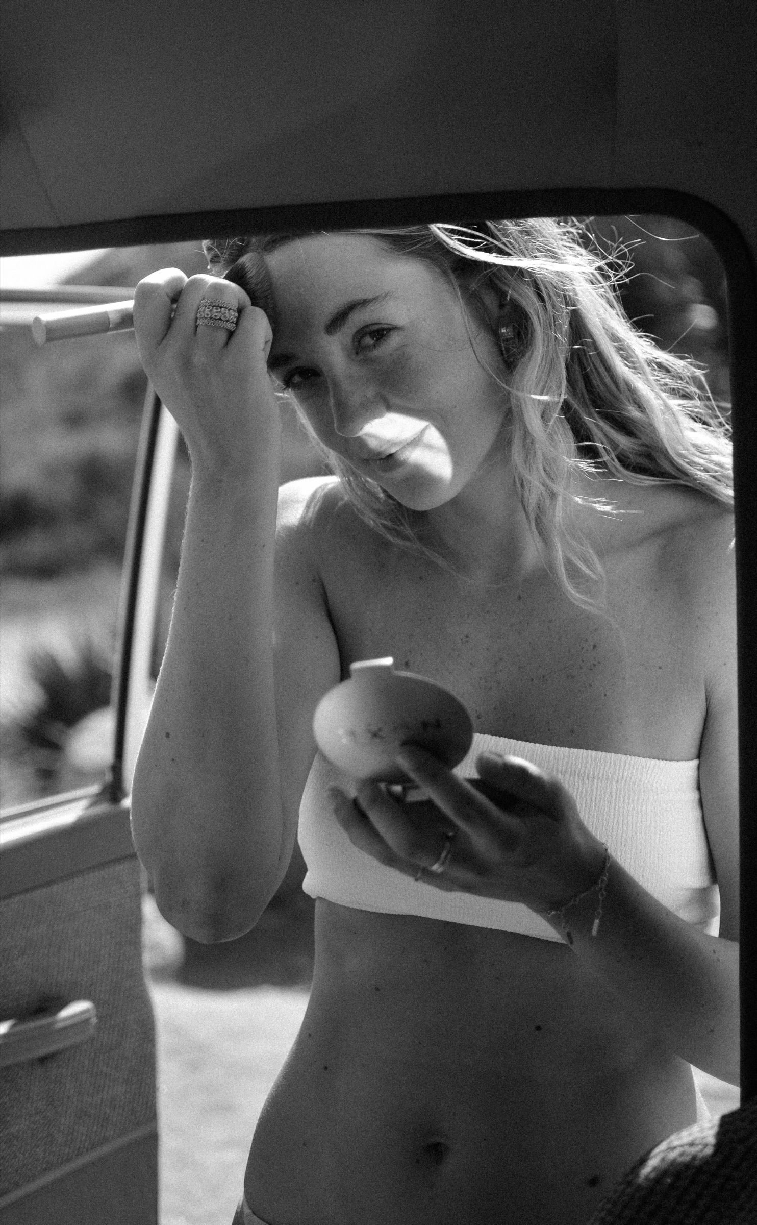 Black-and-white view through a car window of a woman in a bandeau top holding a compact mirror while fixing her hair in bright sun