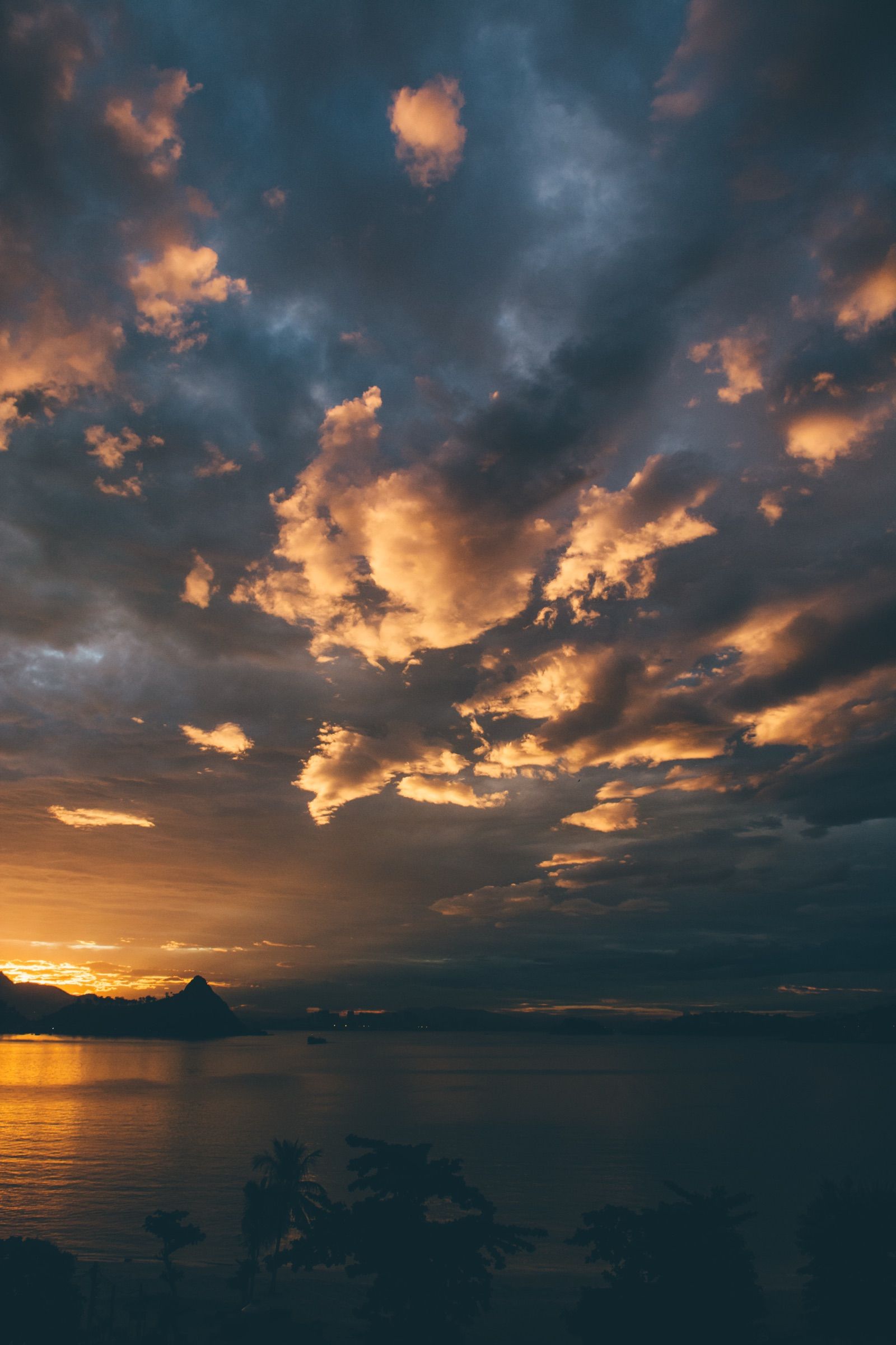 Scattered clouds burning orange at sunset over a dark sea, with a silhouetted headland and palm trees below