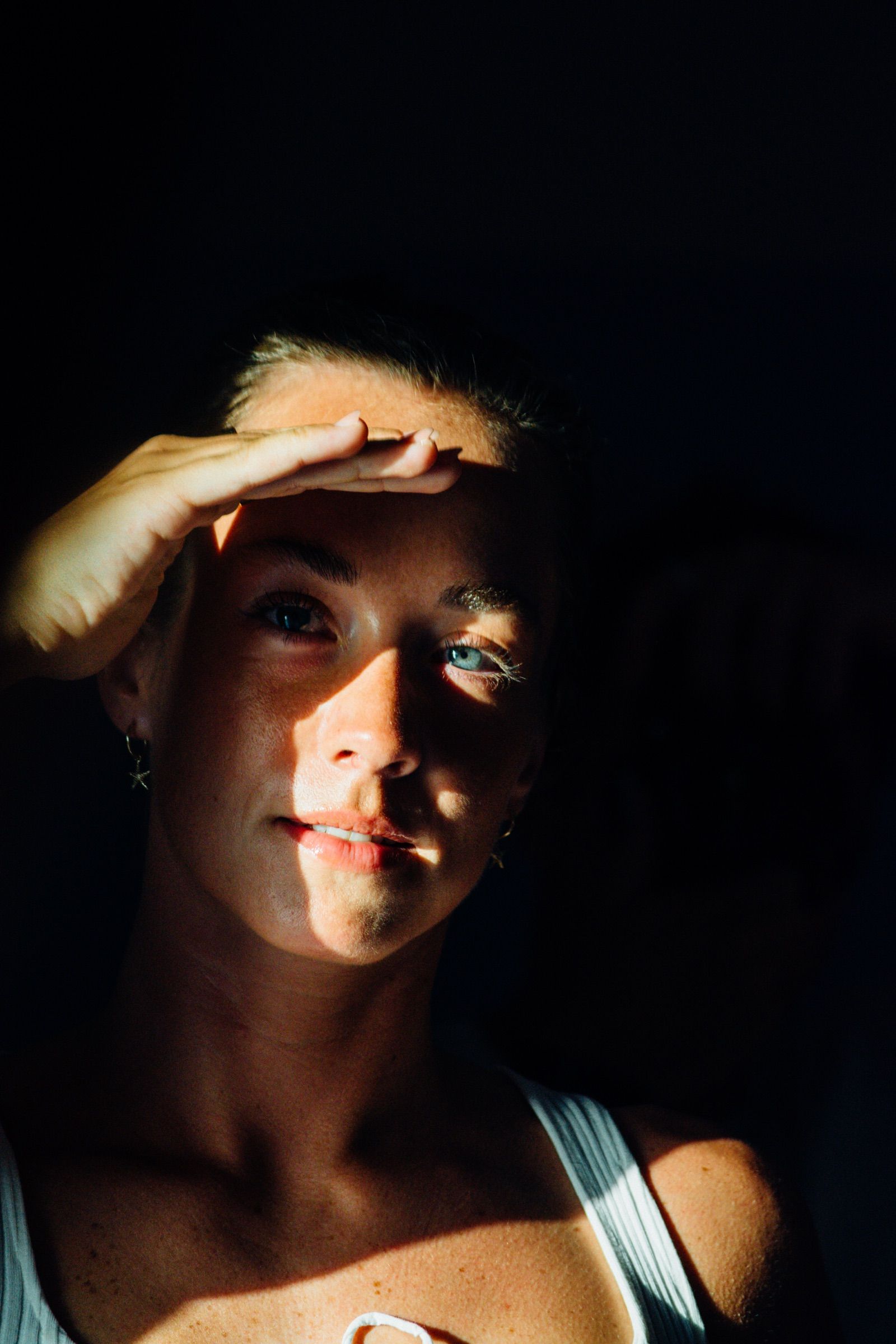 Woman shielding her eyes with one hand as a hard wedge of sunlight cuts across her face and blue eye in deep shadow