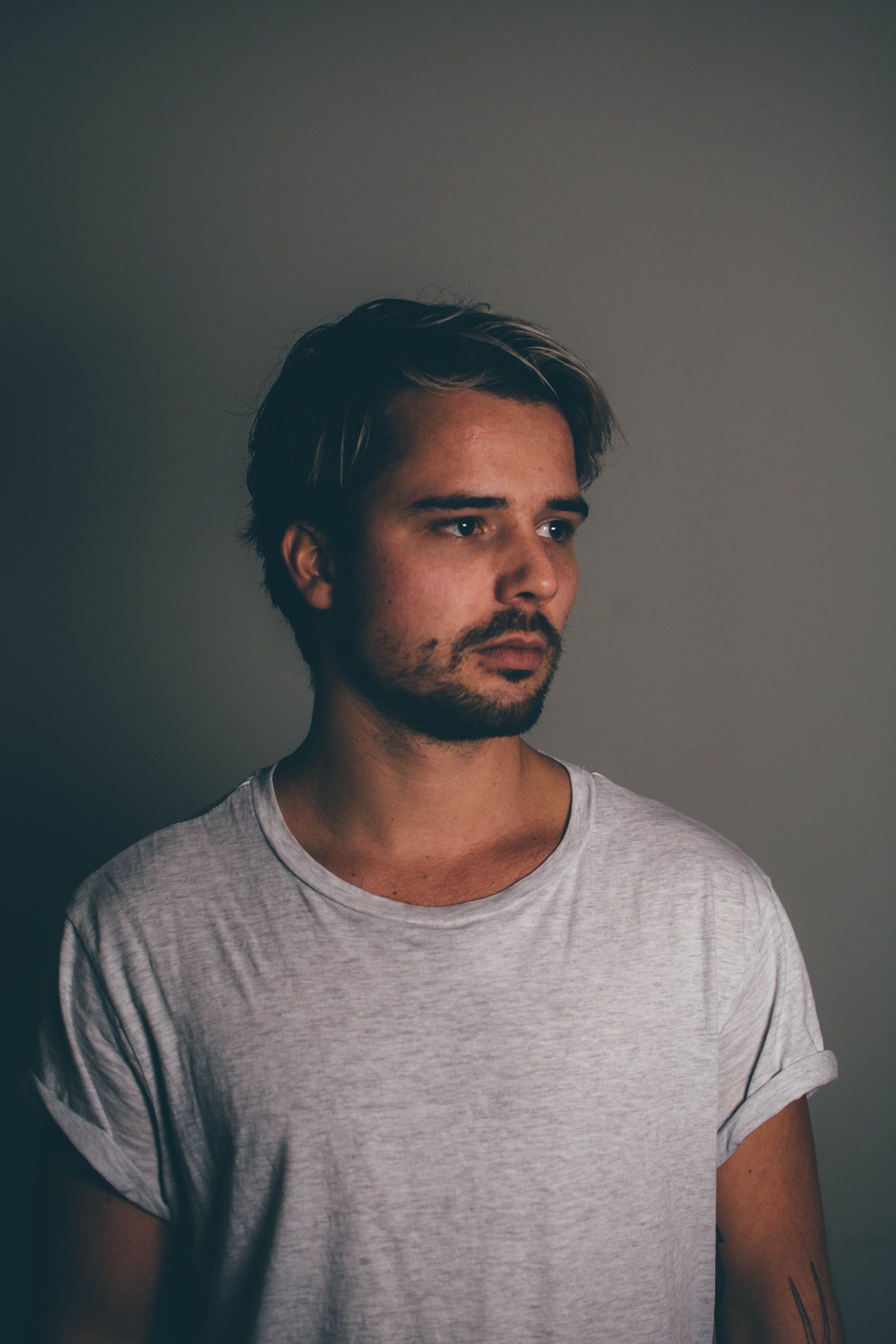 Studio portrait of a man with swept-back blond hair and a beard in a pale t-shirt glancing off-frame against a grey wall