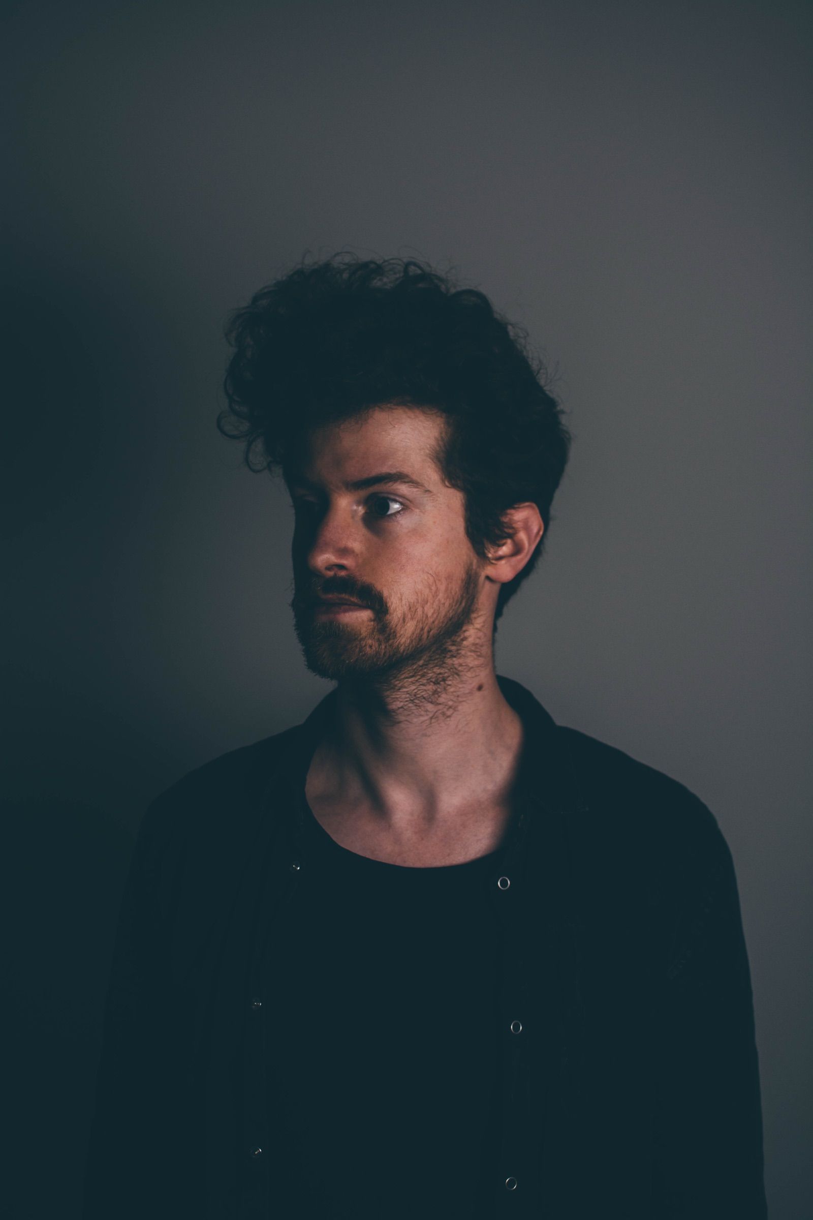 Low-key portrait of a man with dark curly hair and a beard looking sideways in dim grey light