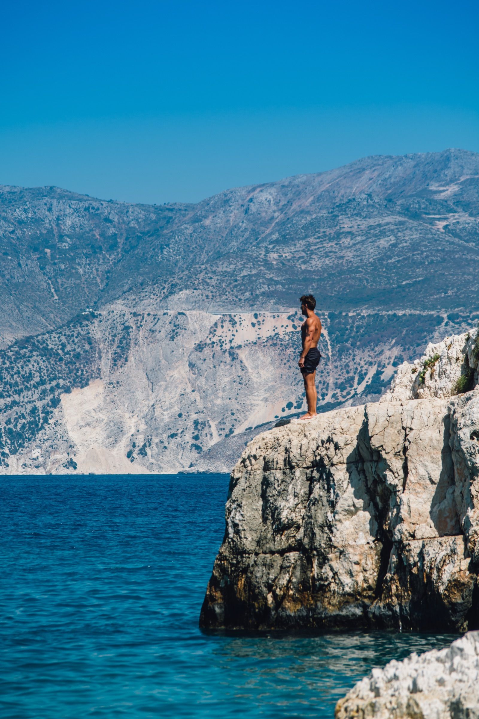 Man in swim shorts standing on a white limestone outcrop, gazing across deep blue water to bare sunbaked mountains