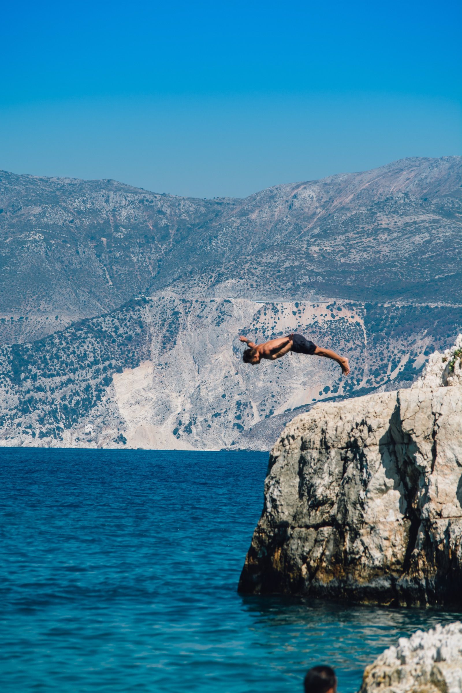 Swimmer diving horizontally from a rocky ledge toward deep blue sea against a backdrop of arid cliffs