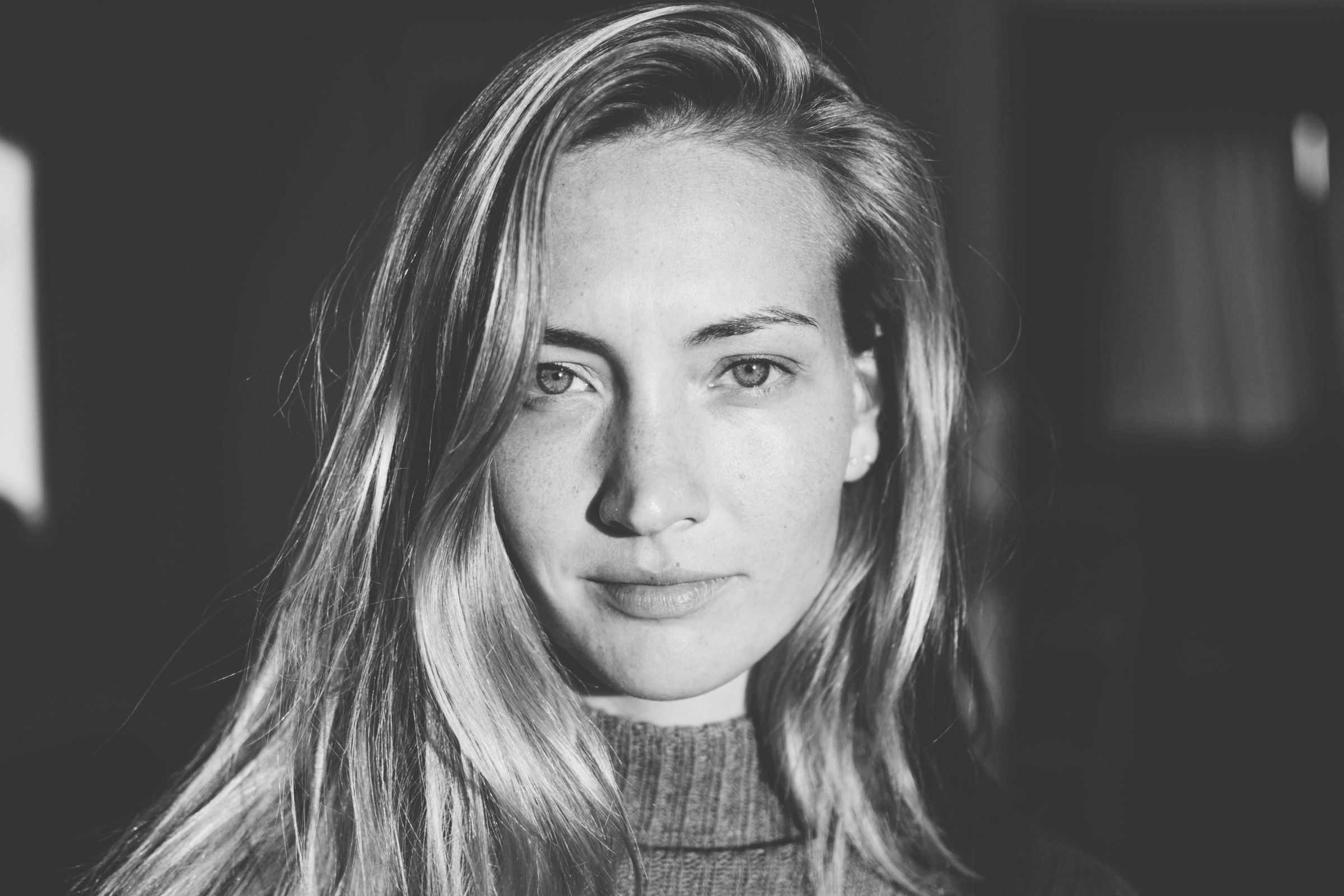 Black-and-white close-up of a woman with long fair hair meeting the camera’s gaze, half her face lit by window light