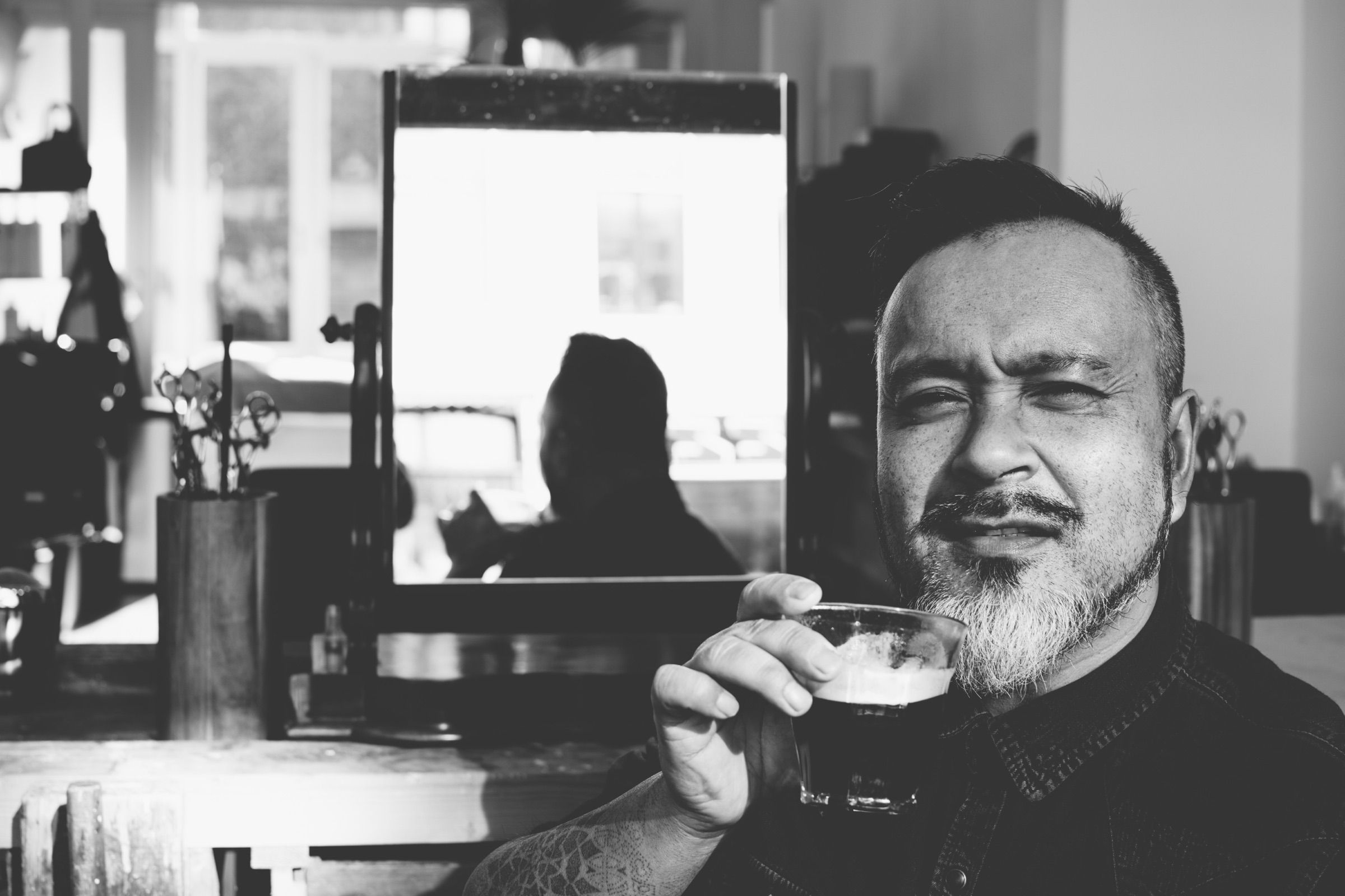 Black-and-white portrait of a squinting man with a grey-flecked beard holding up a small foam-topped glass in a sunlit barbershop