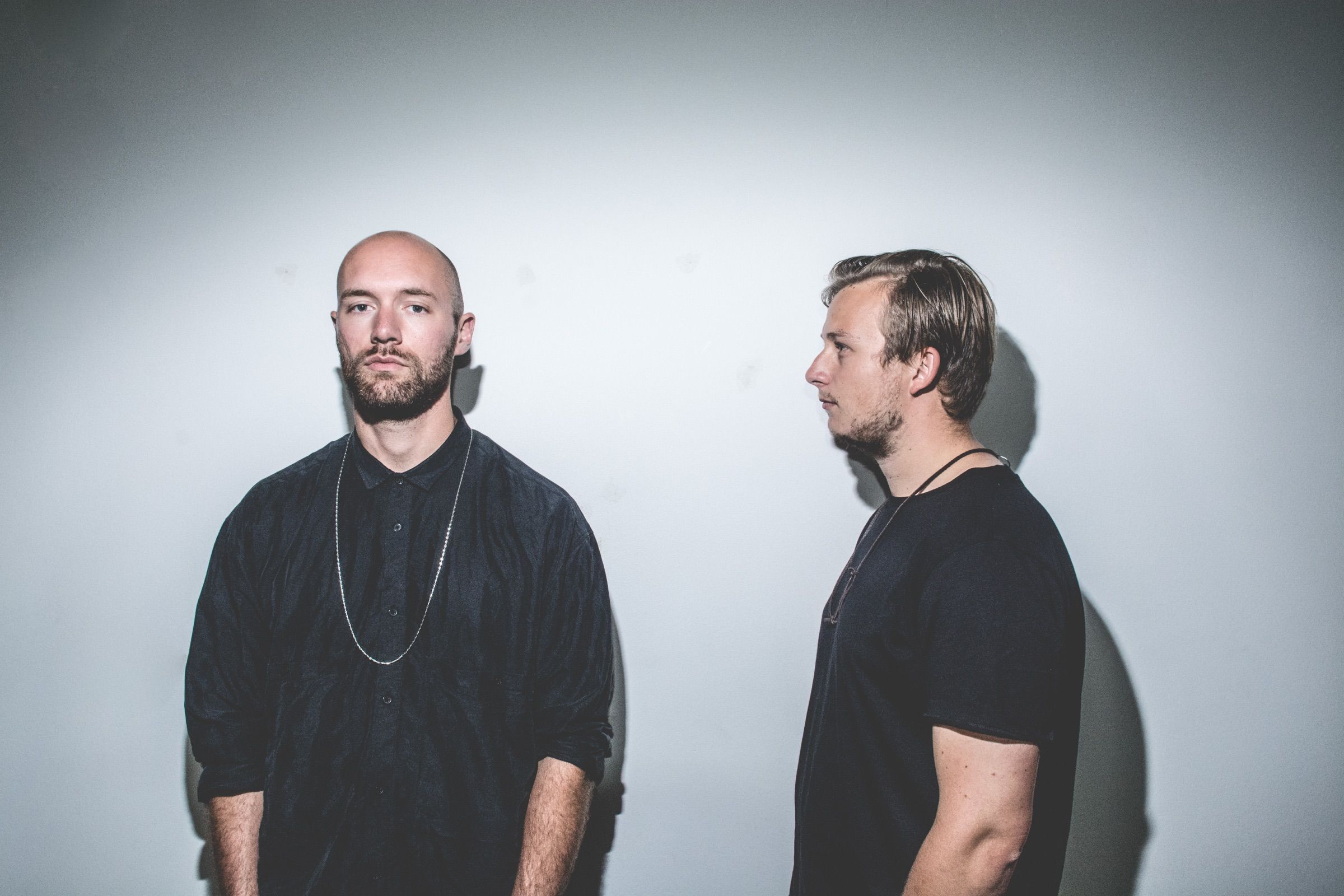 Two men in dark clothes against a flash-lit pale wall, one facing the camera in a chain necklace, the other standing in profile