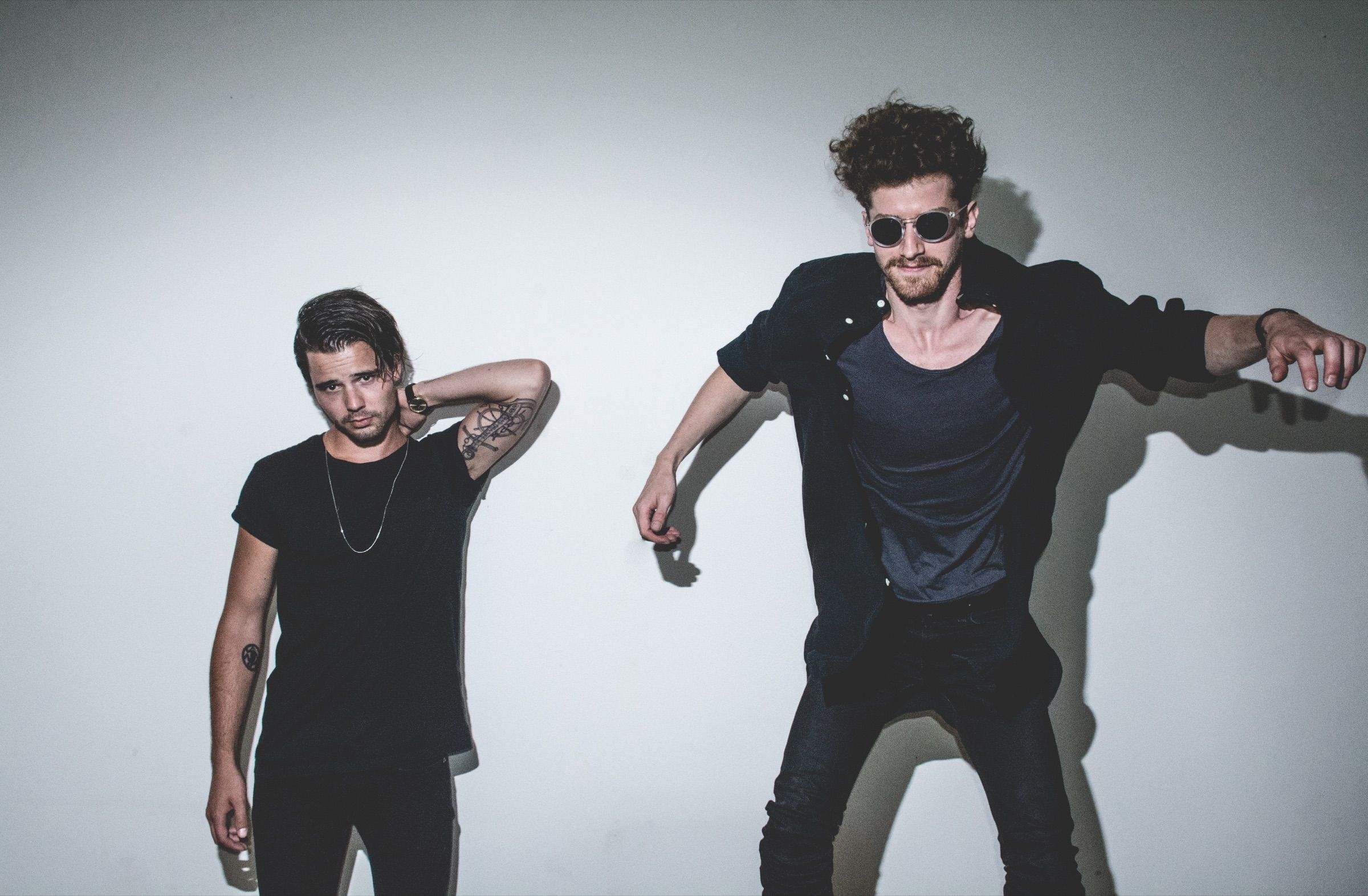 Two band members posing against a white wall, one leaning into frame in round sunglasses with arms flung out