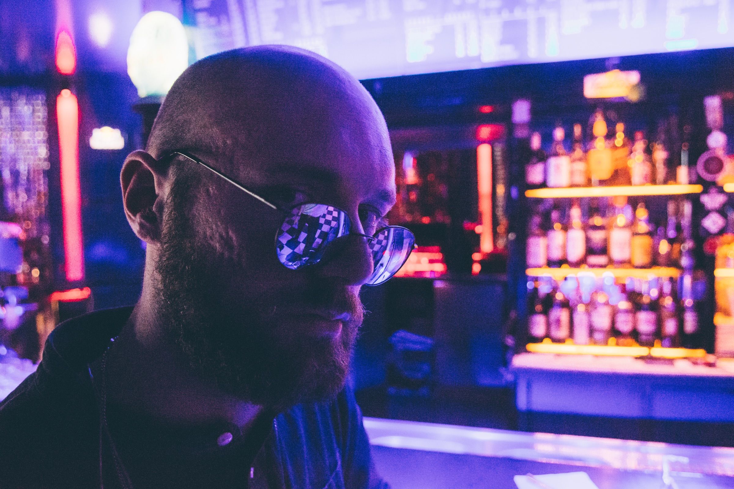 Bald bearded man in round glasses bathed in purple neon light at a bar, shelves of backlit bottles glowing behind