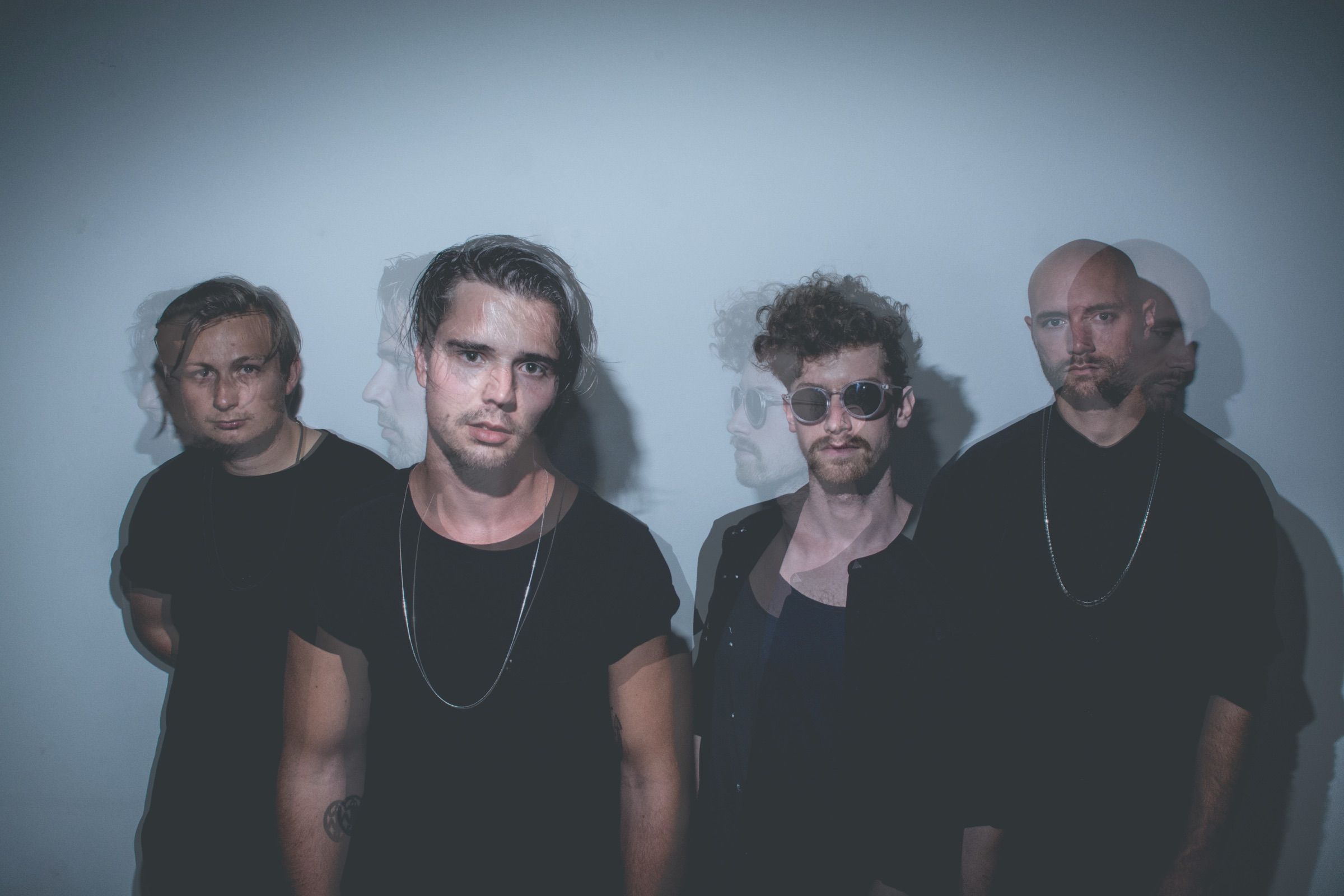 Ghosted double-exposure group portrait of four musicians in dark clothes lined up against a pale wall