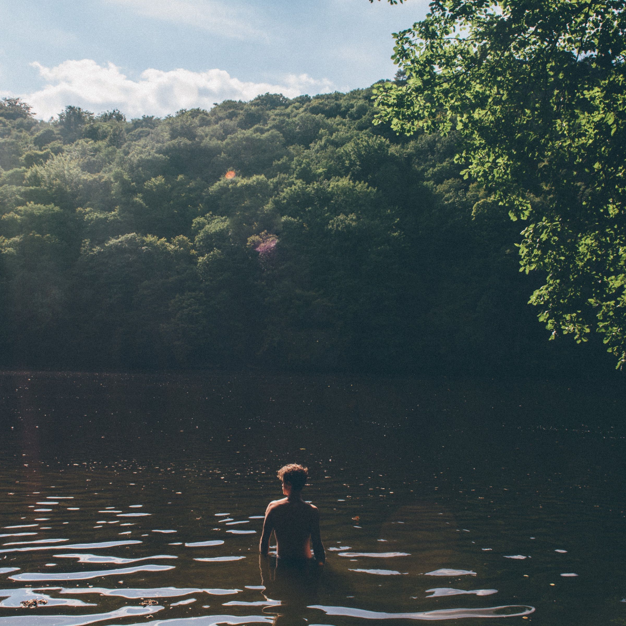 Swimmer standing waist-deep in a dark forest lake, sunlit ripples spreading around him beneath wooded banks