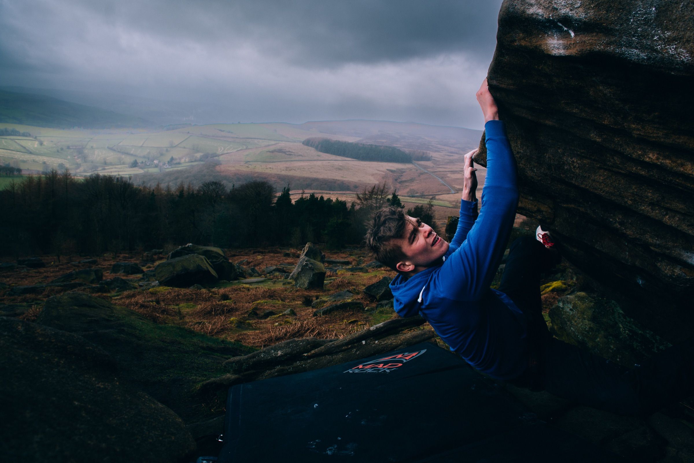 Boulderer in a blue hoodie clinging to an overhanging gritstone block above a crash pad, rain-hazed moorland valley stretching behind