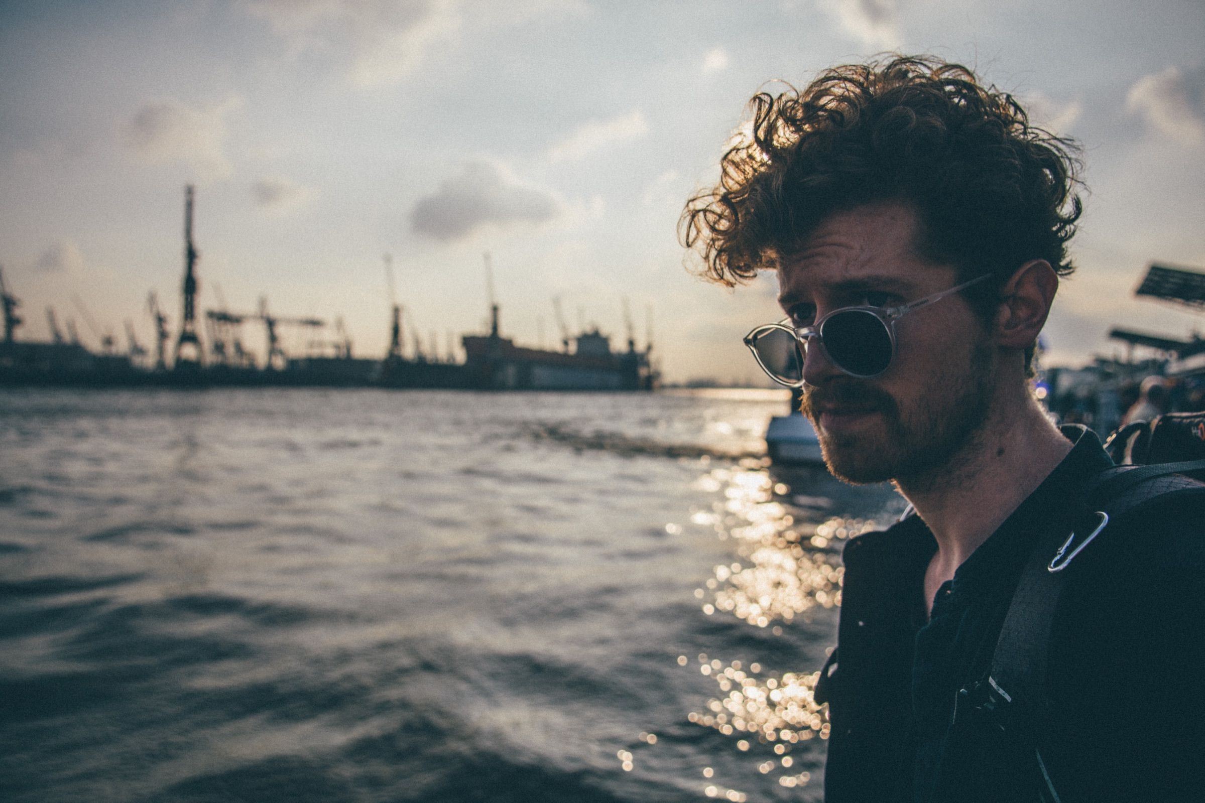 Man with curly hair and clear-framed sunglasses in profile on a harbor ferry, dockyard cranes and glittering water behind him