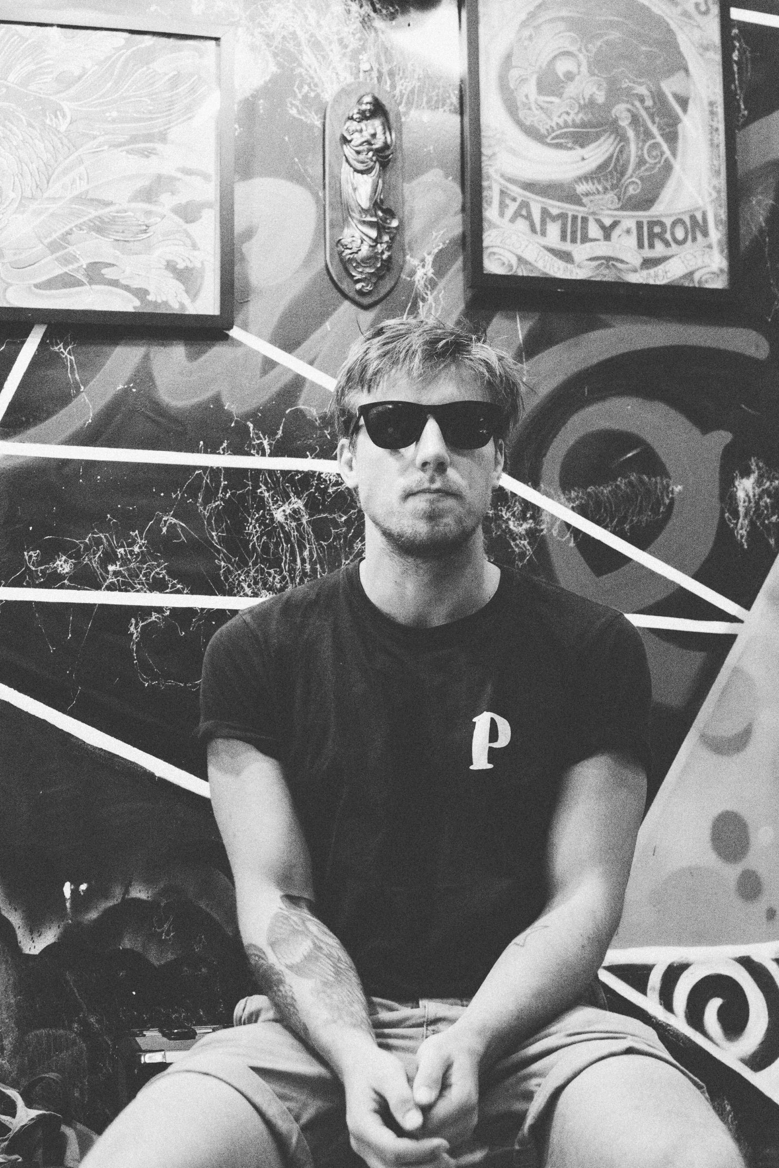 Black-and-white portrait of a man in dark sunglasses sitting with clasped hands before a graffiti-covered wall hung with framed tattoo art