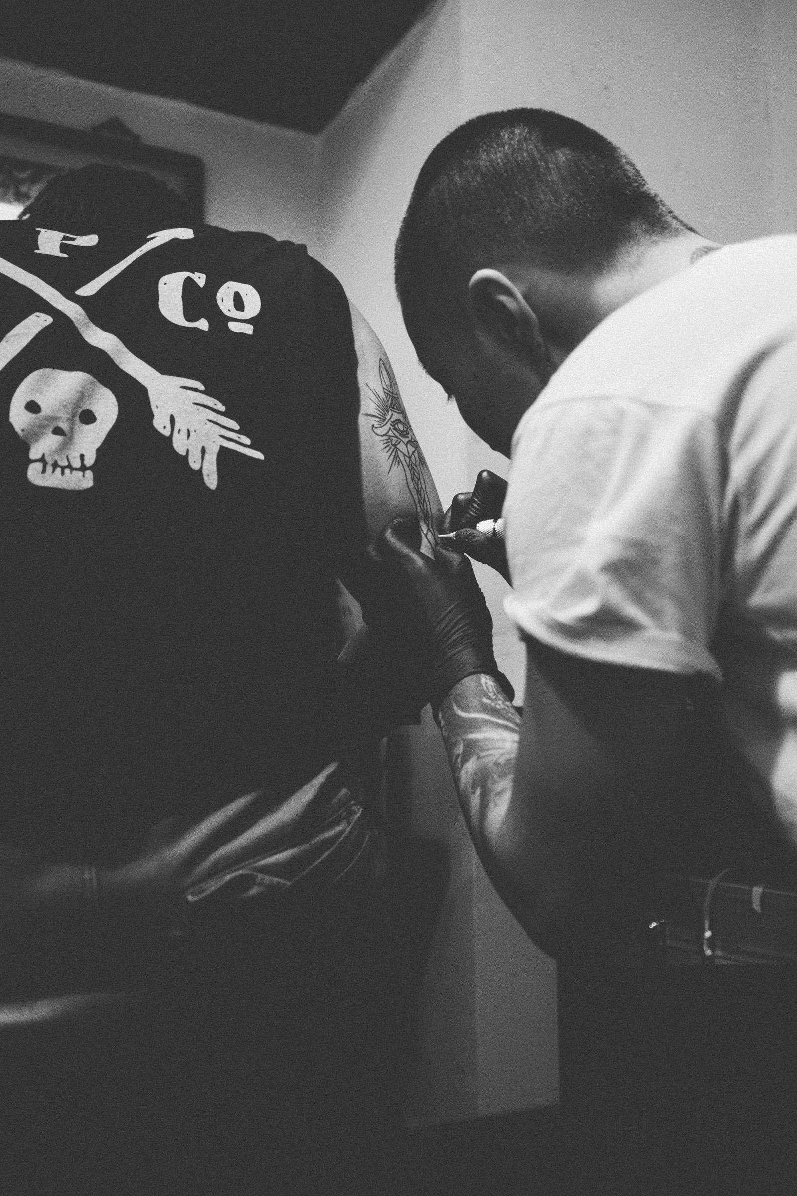 Black-and-white scene of a tattoo artist in black gloves bent close over a client’s shoulder, inking a fresh outline