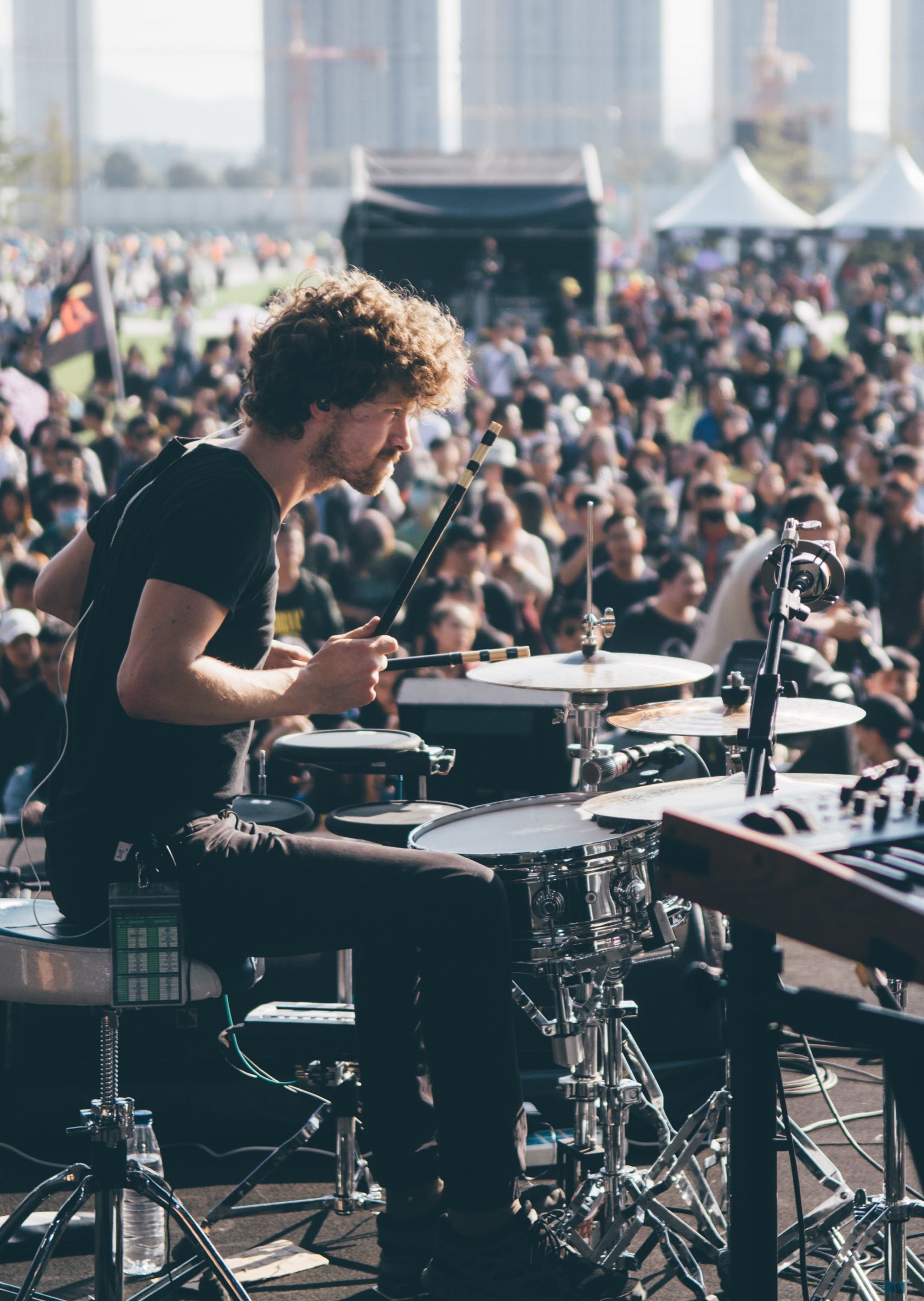 Drummer with curly hair playing side-on at an outdoor festival stage before a huge crowd stretching toward city towers