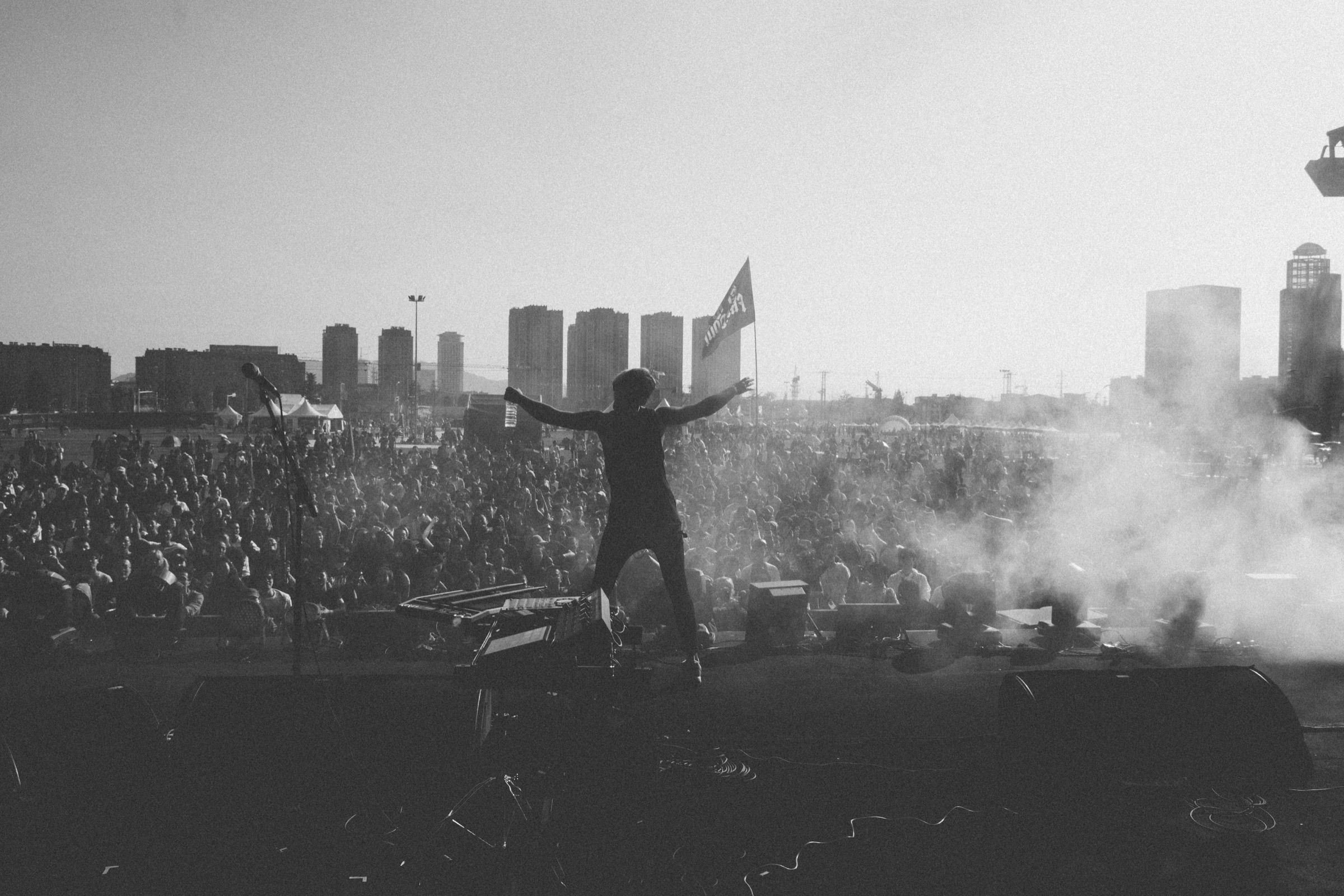 Black-and-white view from behind a performer with arms flung wide at the stage edge before a vast festival crowd and hazy skyline