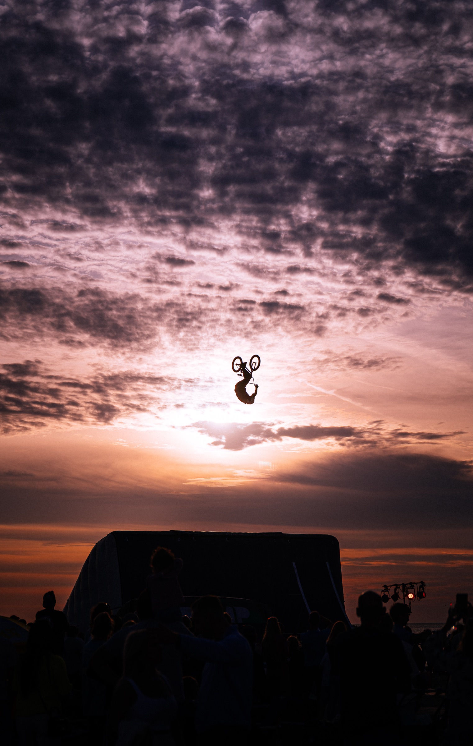 BMX rider silhouetted mid-backflip high against a glowing purple and orange sunset sky above a darkened crowd and landing ramp