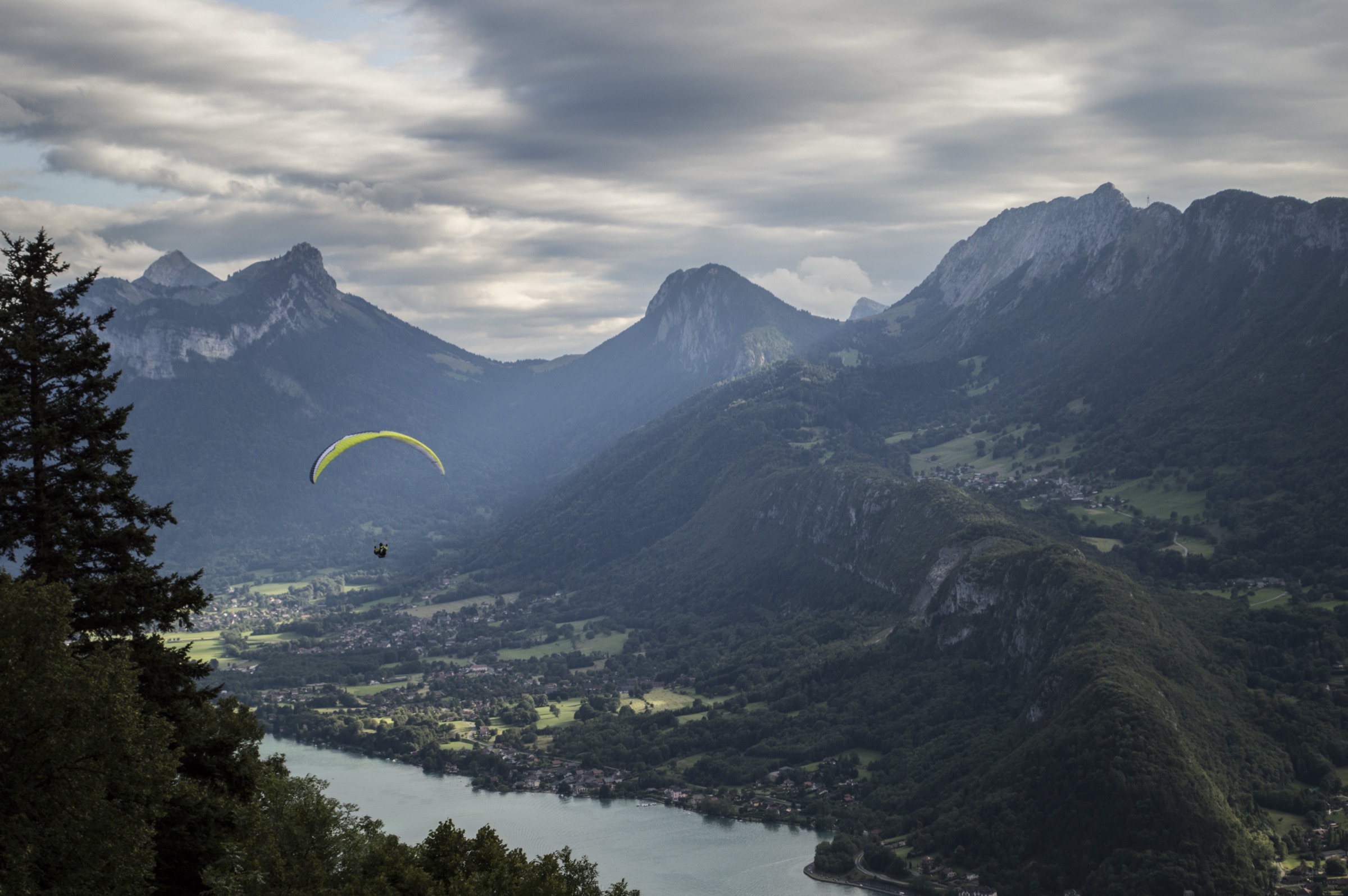 Paraglider with a yellow wing soaring above an alpine lake valley ringed by mountains under moody clouds
