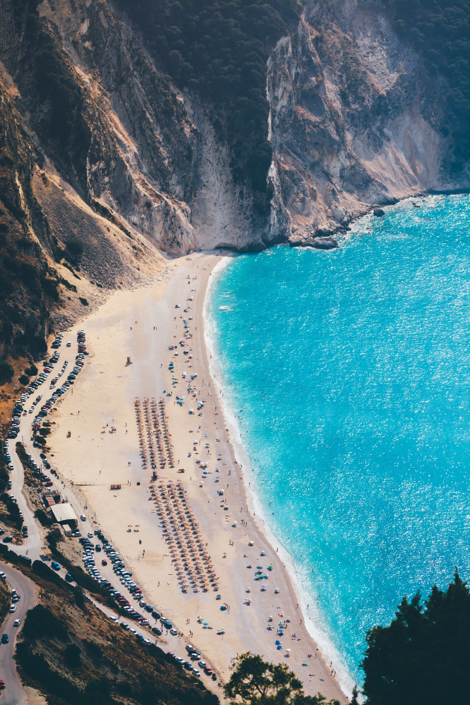 Aerial view of a white pebble beach with rows of umbrellas curving along turquoise water beneath steep cliffs