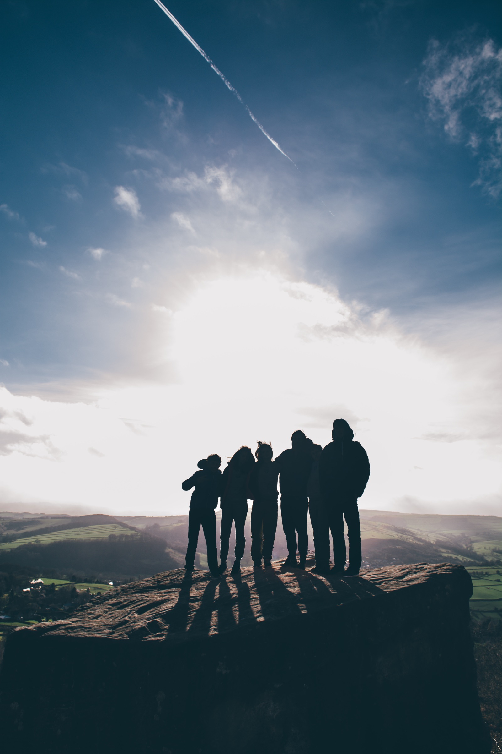 Six friends silhouetted shoulder to shoulder on a rocky outcrop against a bright backlit sky crossed by a contrail