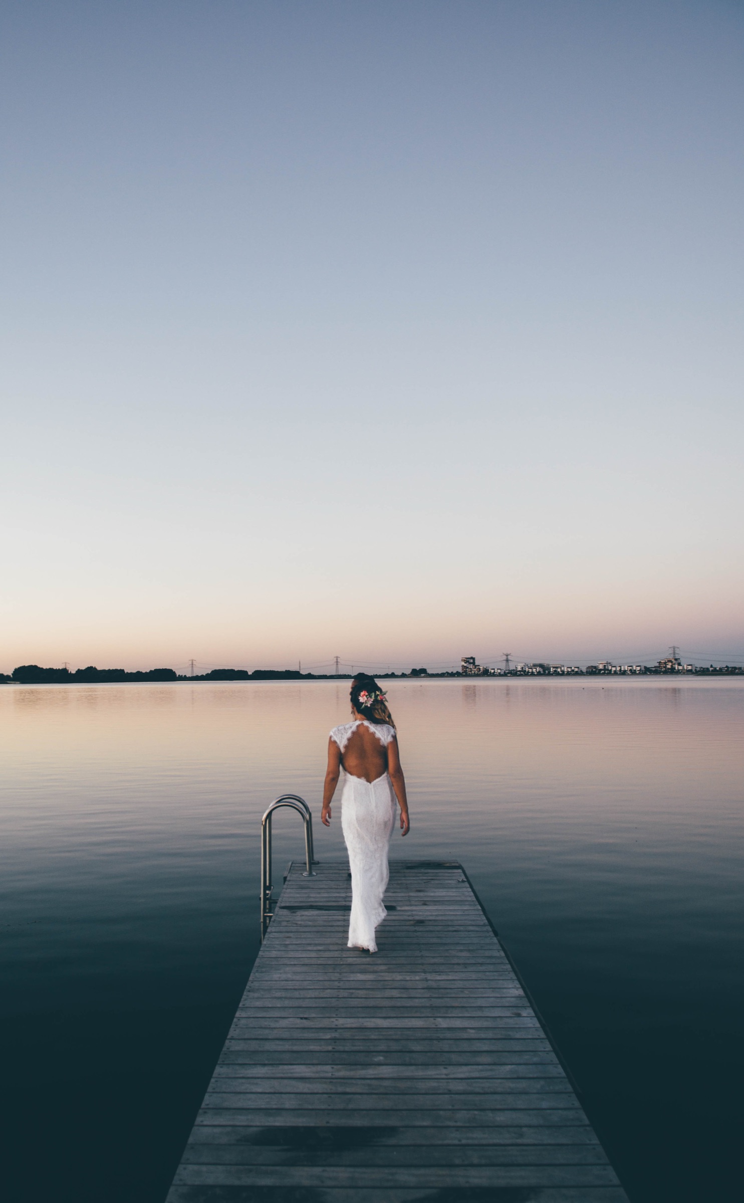 Bride in a backless lace gown walking down a wooden jetty over a mirror-calm lake at dusk