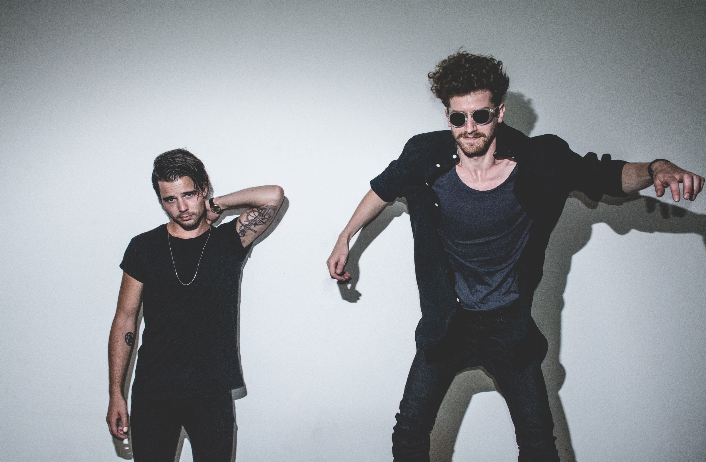 Two band members posing against a white wall, one leaning into frame in round sunglasses with arms flung out