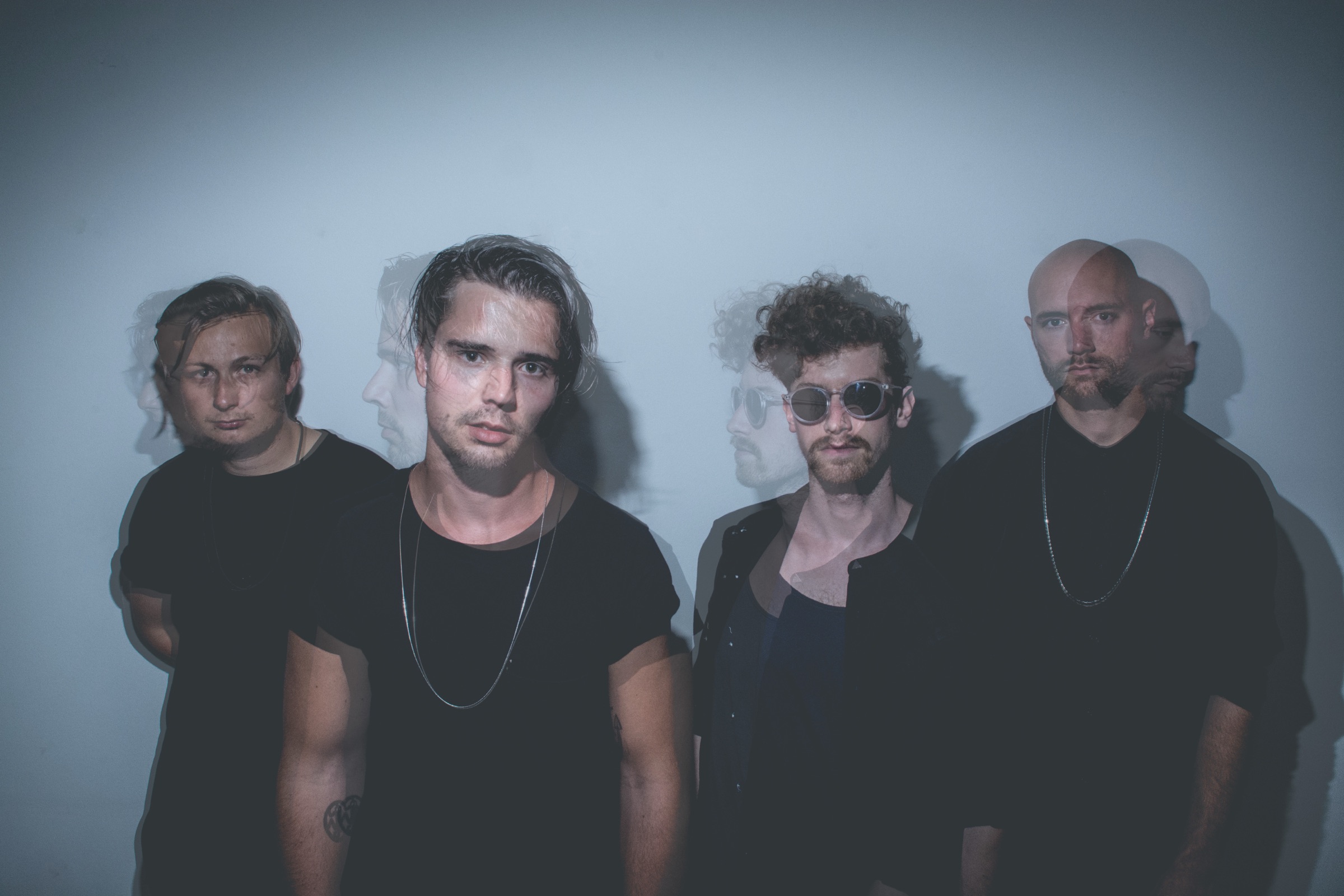 Ghosted double-exposure group portrait of four musicians in dark clothes lined up against a pale wall